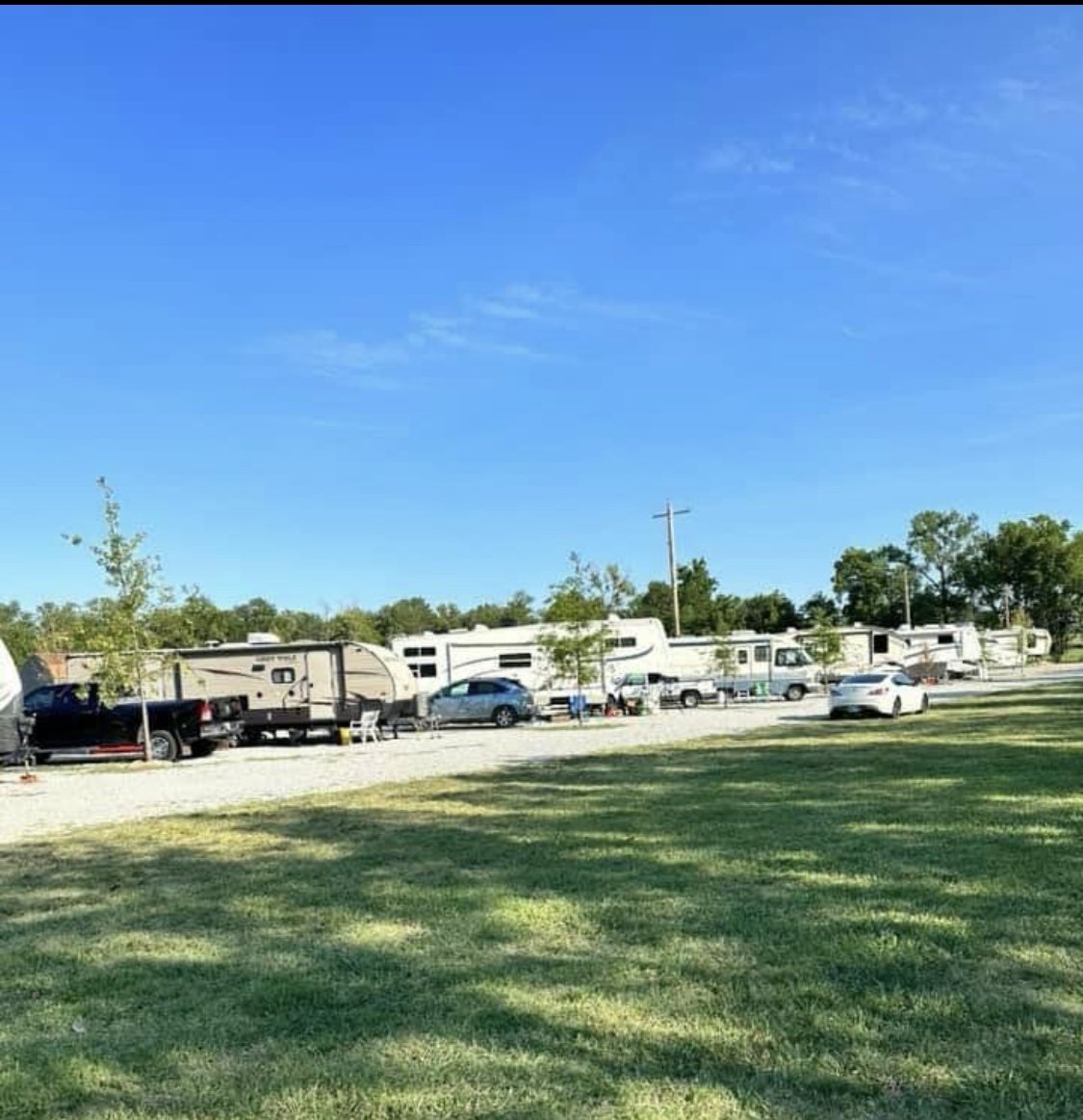 Outdoor camping area with several RVs and trailers parked along a gravel lot, surrounded by greenery and trees under a clear blue sky.
