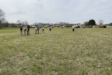 A group of people standing on a large grassy field under a cloudy sky, with some vehicles and trees in the background.