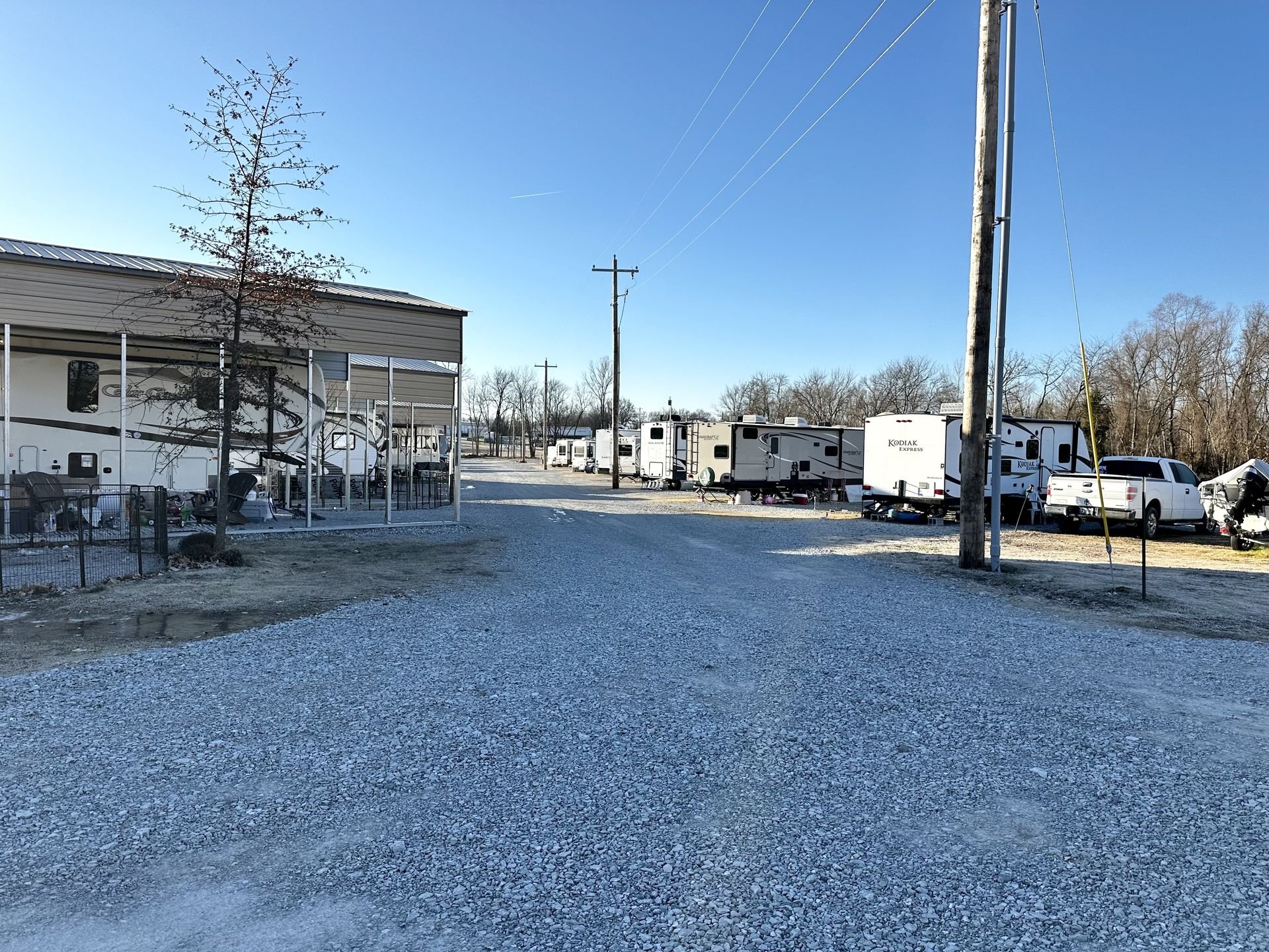Gravel road with parked RVs and trailers beside a metal-roofed structure, surrounded by leafless trees on a clear day.