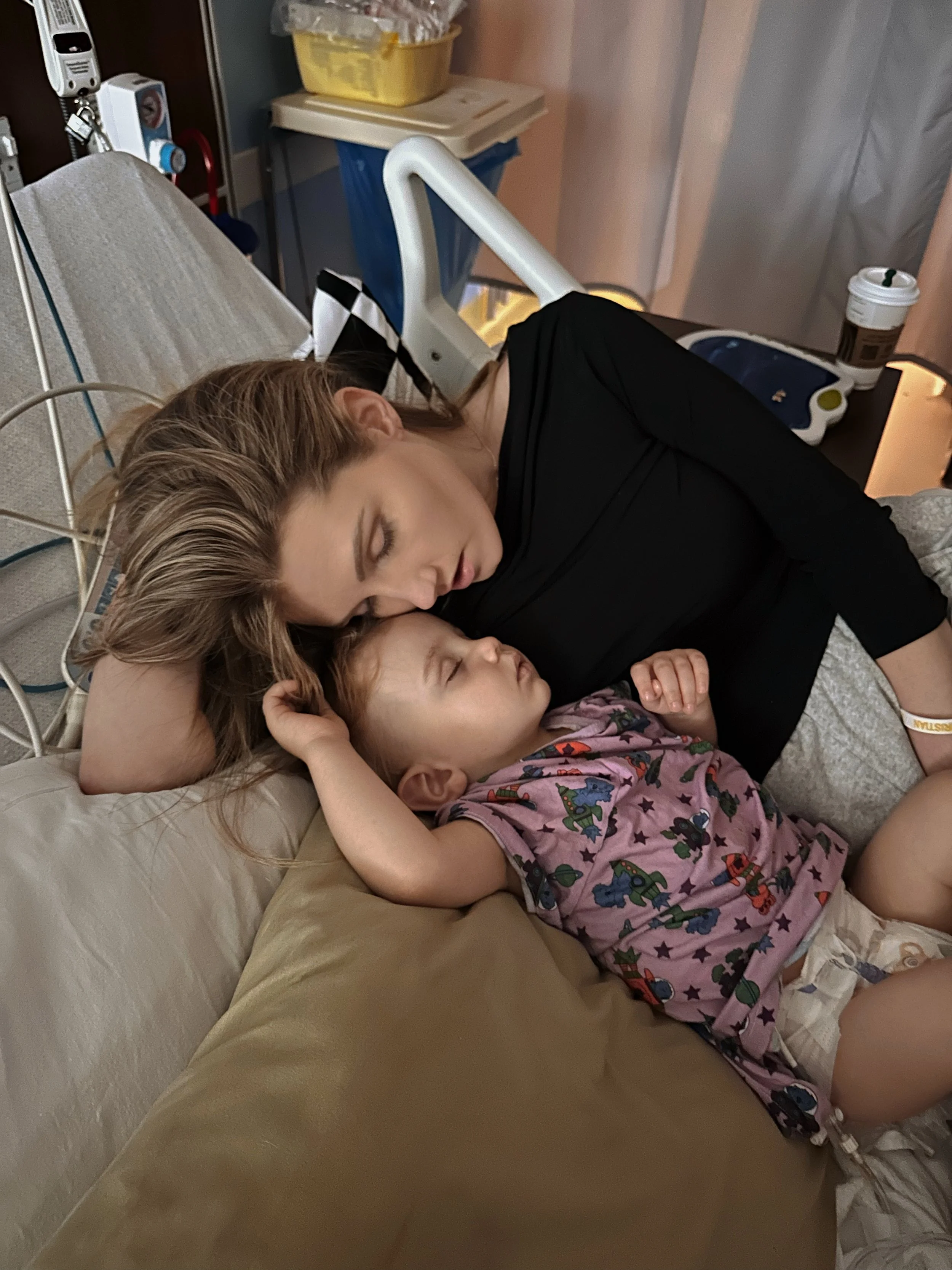 A woman and a child sleeping together on a hospital bed. The woman is resting her head on a pillow, while the child is wearing a printed shirt and diaper. Medical equipment and a cup are visible in the background.