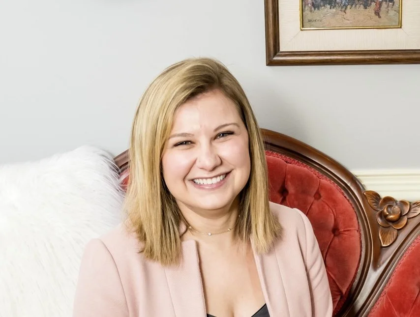 A smiling young woman with shoulder-length blonde hair, wearing a pink blazer, sitting on a red velvet antique chair in a room with framed artwork on the wall.
