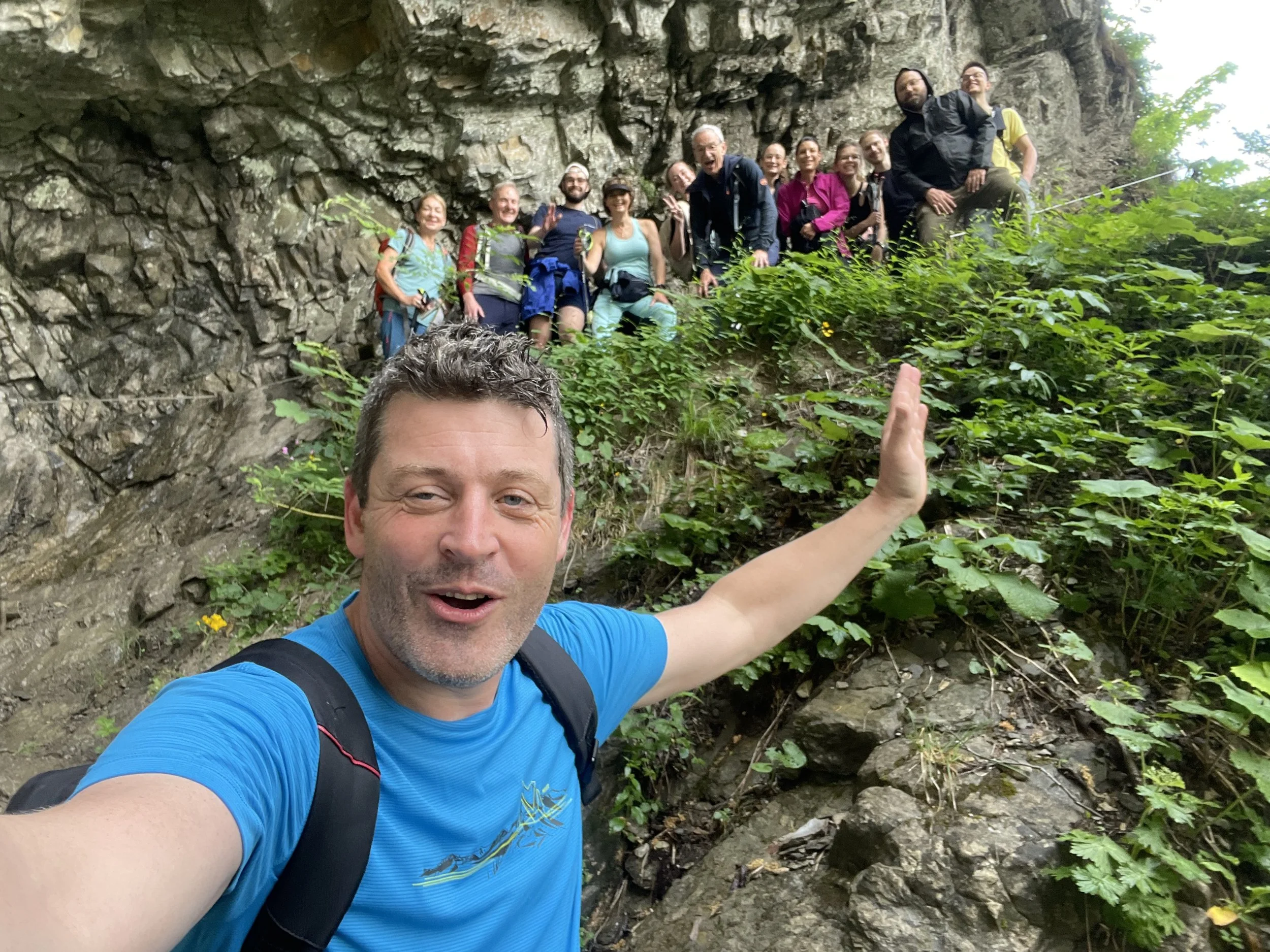 Ein Mann macht ein Selfie vor einer Gruppe von Wanderern auf einer felsigen, grünen Steilwand im Freien.