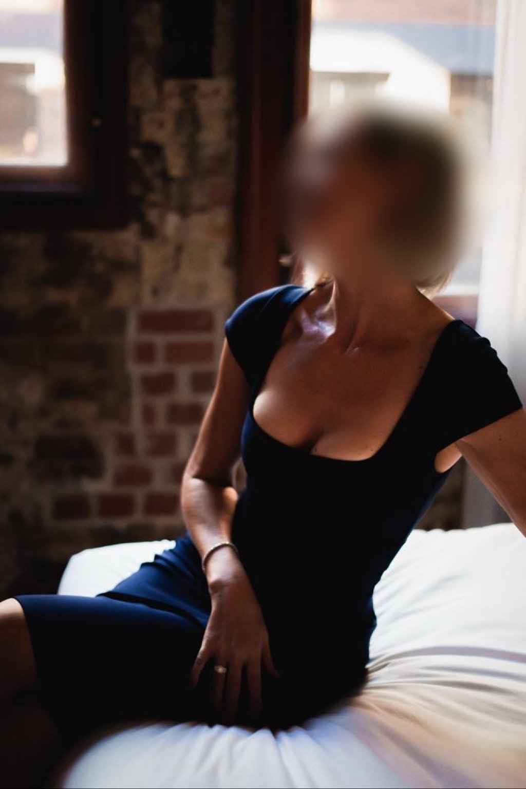 Person wearing a black dress sitting on a white bed in a room with brick walls.