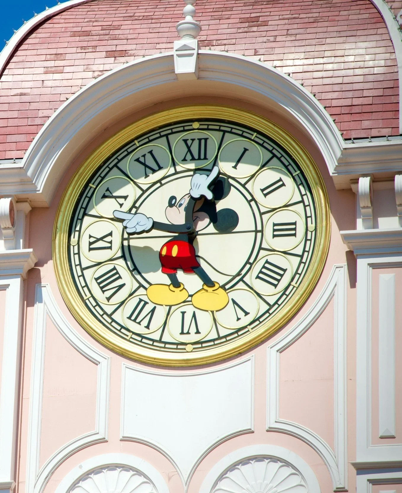 Clock featuring Mickey Mouse pointing at the time, with Roman numerals on the face, set against a pink building facade.
