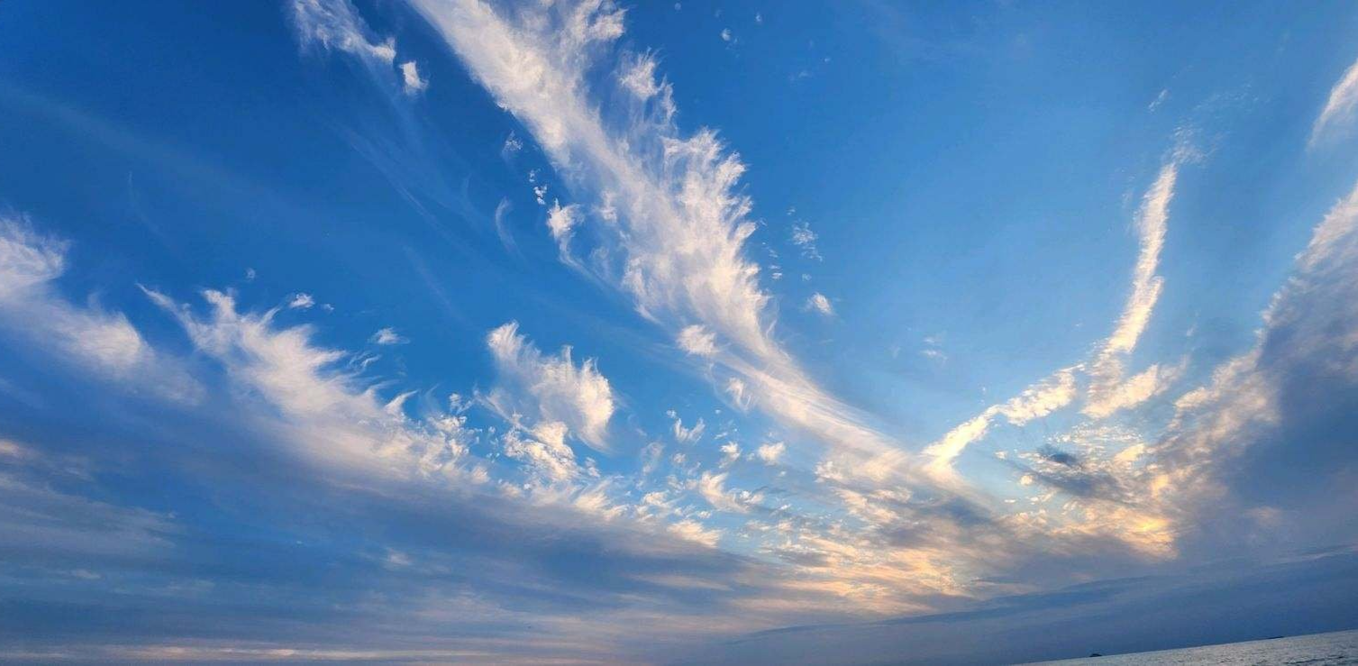Blue sky with scattered white clouds over the ocean with a faint hint of sunset in the distance.