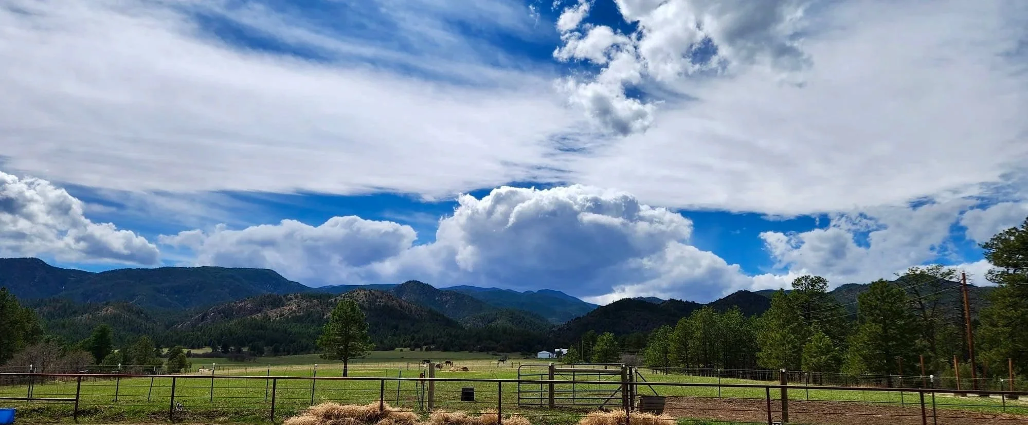 Scenic view of mountains with partly cloudy sky, green fields, trees, and a fenced area in the foreground.