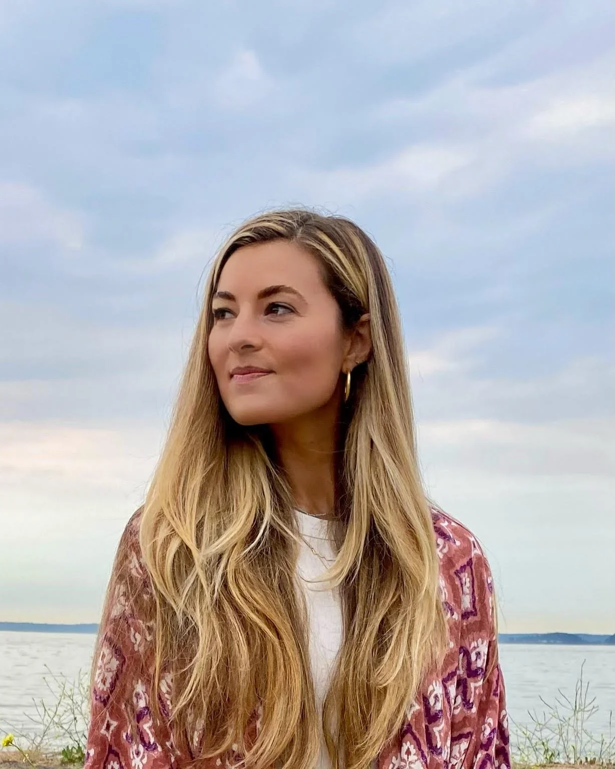 A woman with long blonde hair and gold hoop earrings standing outdoors near a body of water under a cloudy sky.