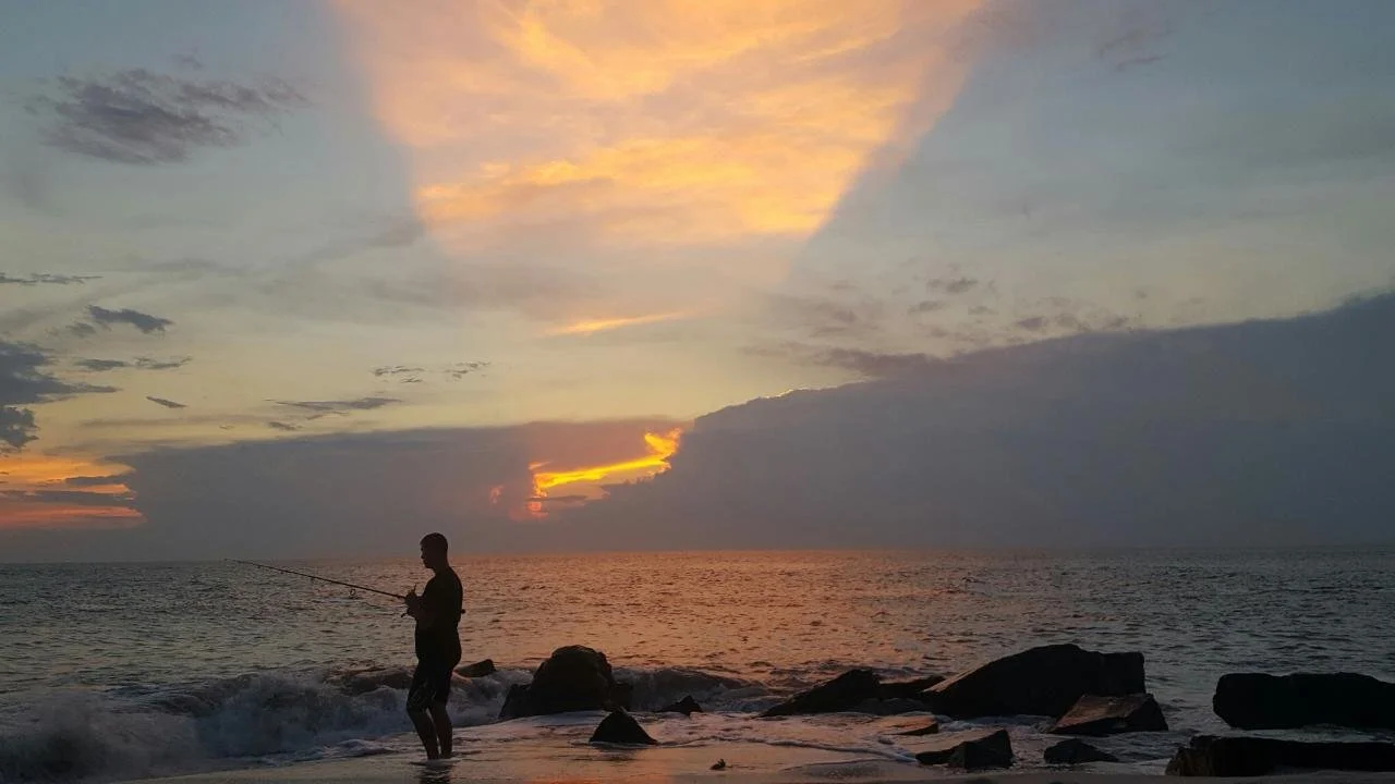 Person fishing on the rocky beach at sunset with colorful sky and calm ocean.