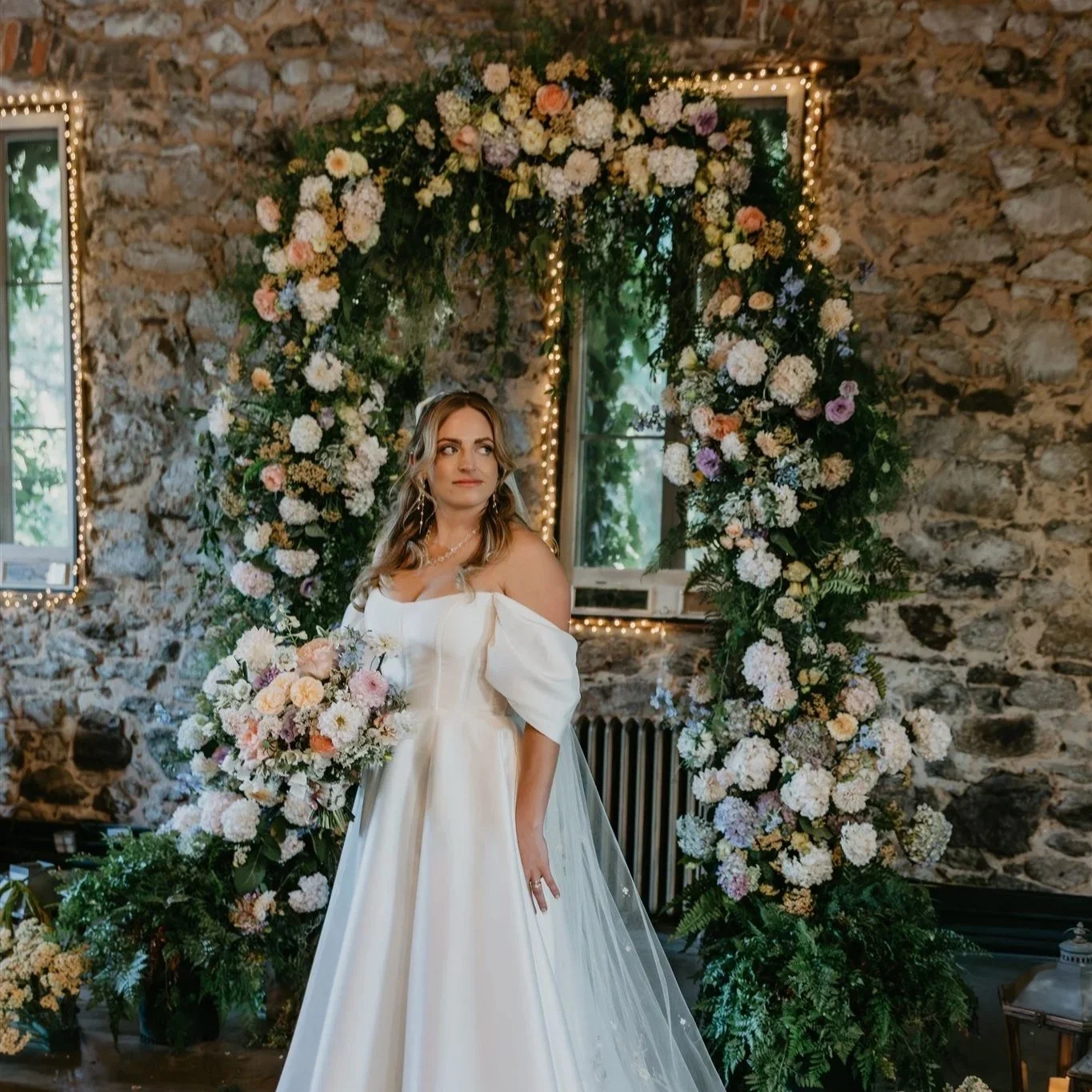 A bride standing in front of a floral arch with white, pink, and purple flowers, inside a rustic stone-walled room with windows and string lights.