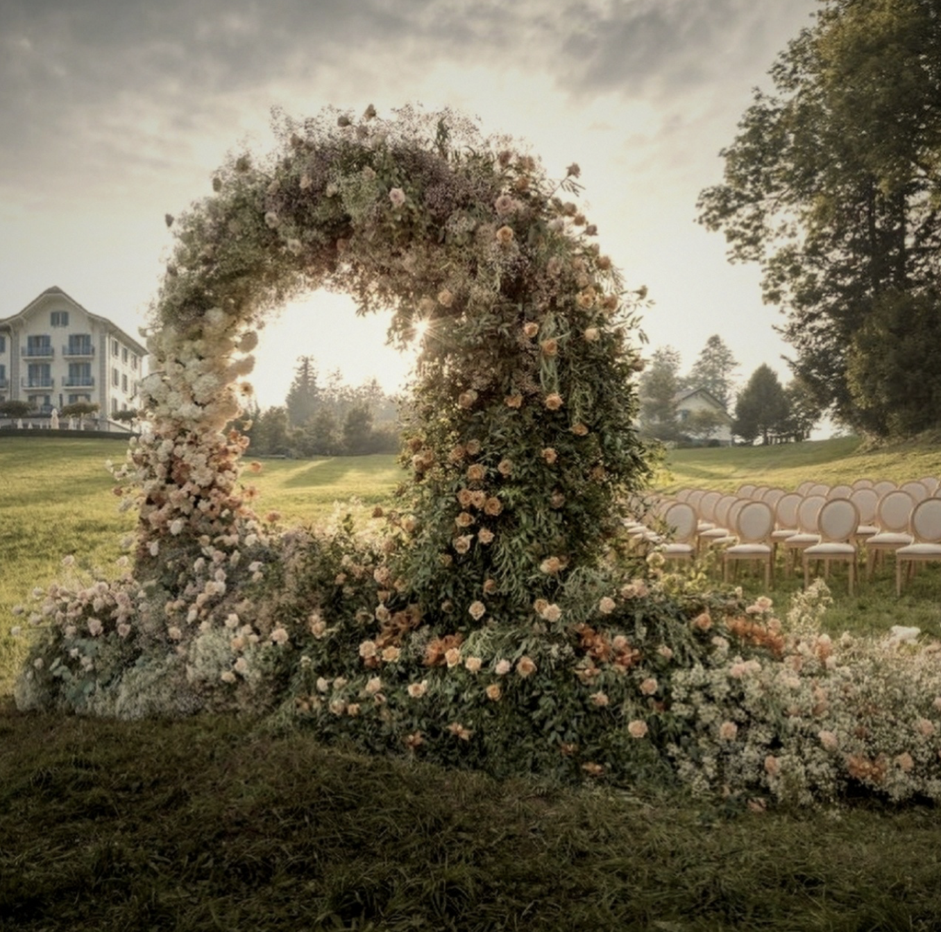 A floral wedding arch decorated with light pink, white, and peach-colored flowers, set outdoors during daytime on a grassy field with empty chairs lined up for guests.