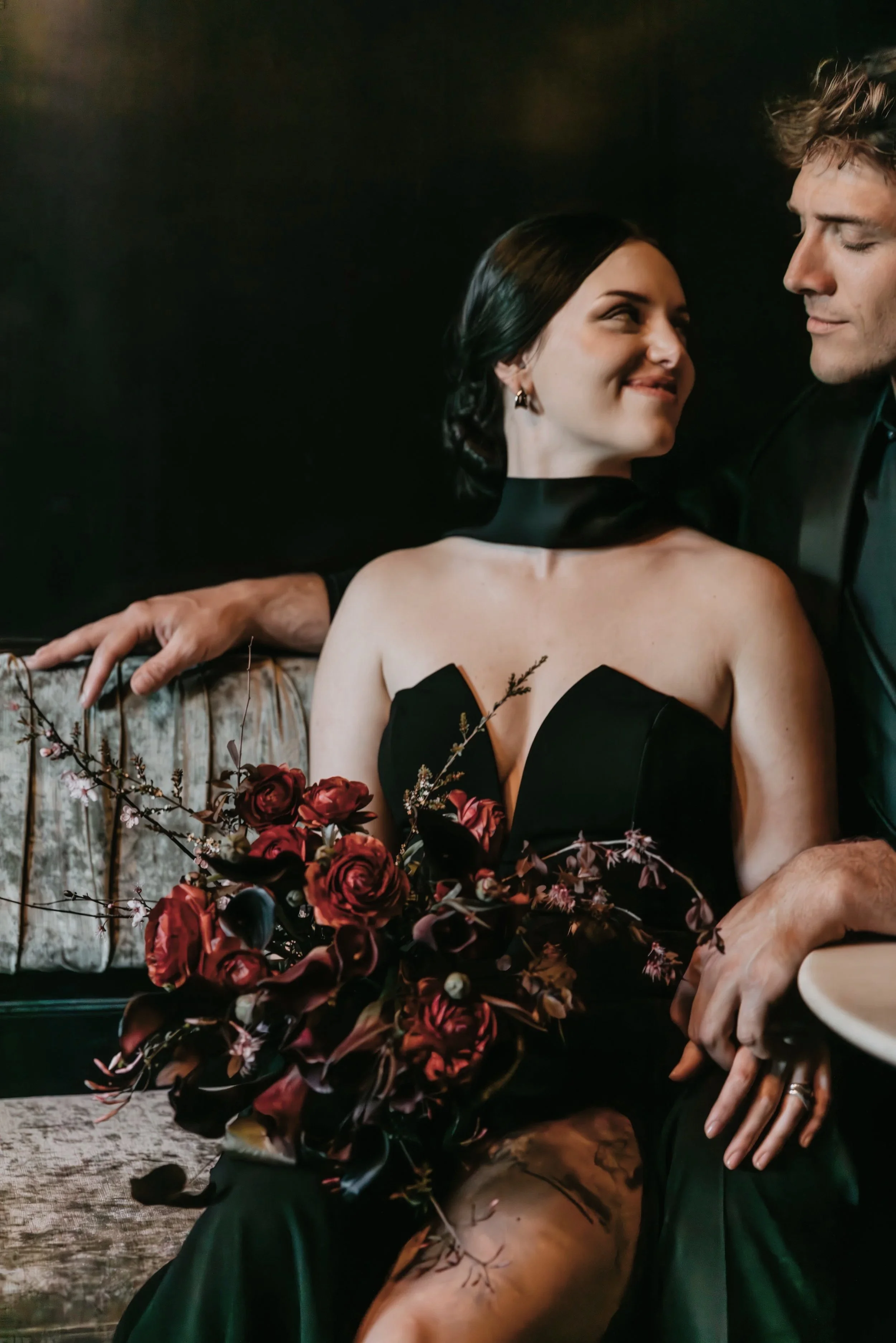 A woman and man sitting close together, with the woman holding a bouquet of dark red flowers. The woman is smiling, wearing a black strapless dress and earrings, while the man has his eyes closed and is dressed in a dark suit. They are sitting on a vintage-style sofa with a dark background.