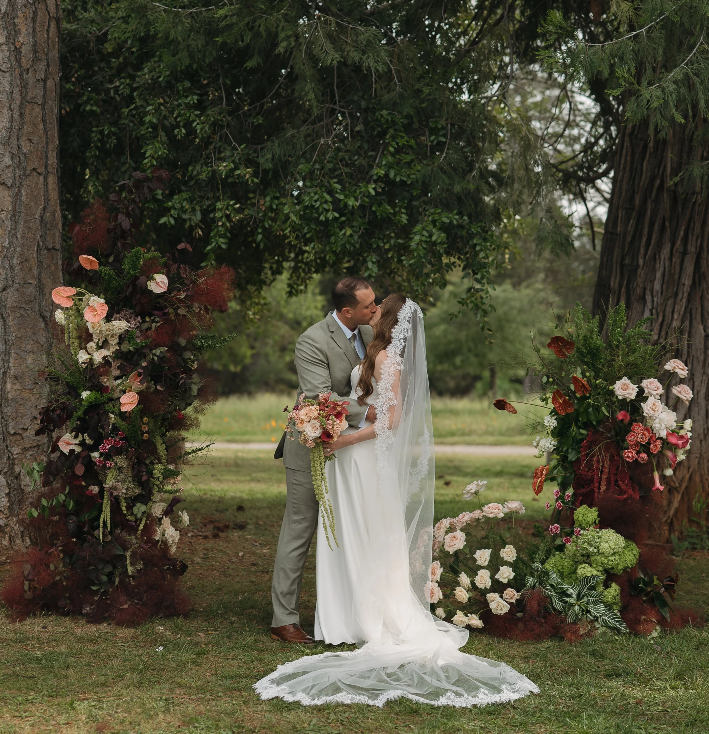 A bride and groom kissing under a floral wedding arch in an outdoor setting in the Sierra Nevada, surrounded by trees and greenery.