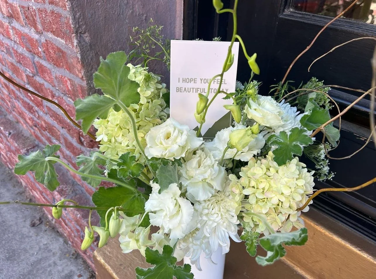 A white bouquet of flowers with a greeting card that says, 'I hope you feel beautiful today,' placed next to a brick wall and a black window frame. Flower delivery in Nevada City and Grass Valley