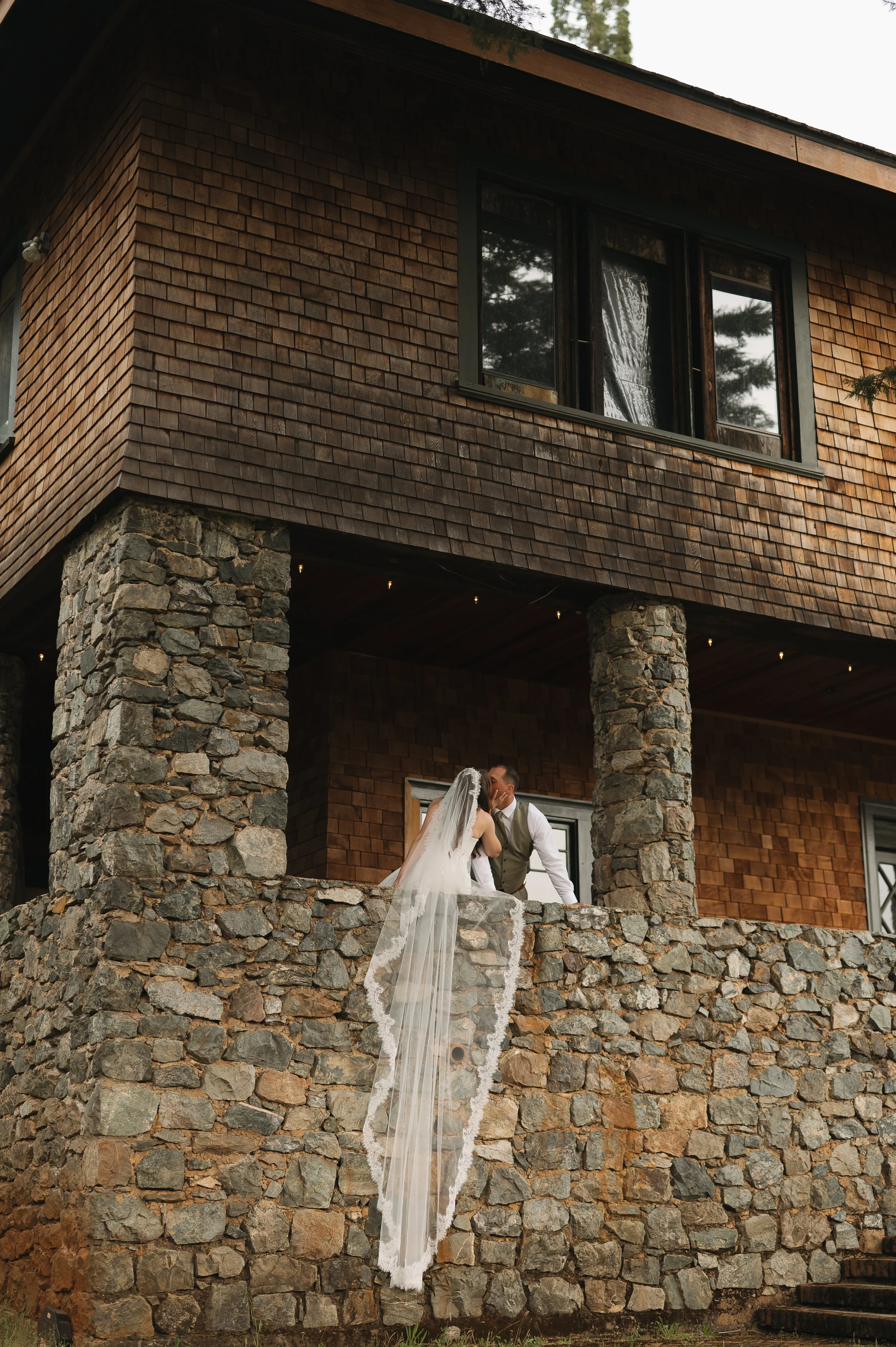 A bride and groom sharing a kiss on a stone balcony of a rustic wooden house with large windows, surrounded by tall trees in the background.