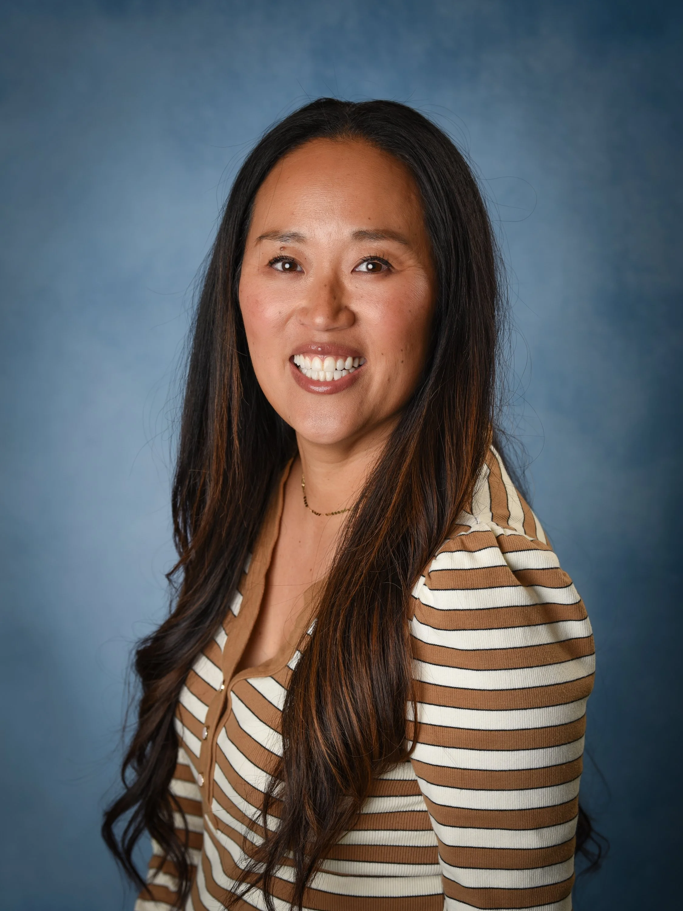 Smiling woman with long straight brown hair, wearing a striped blouse and thin necklace against a blue background.