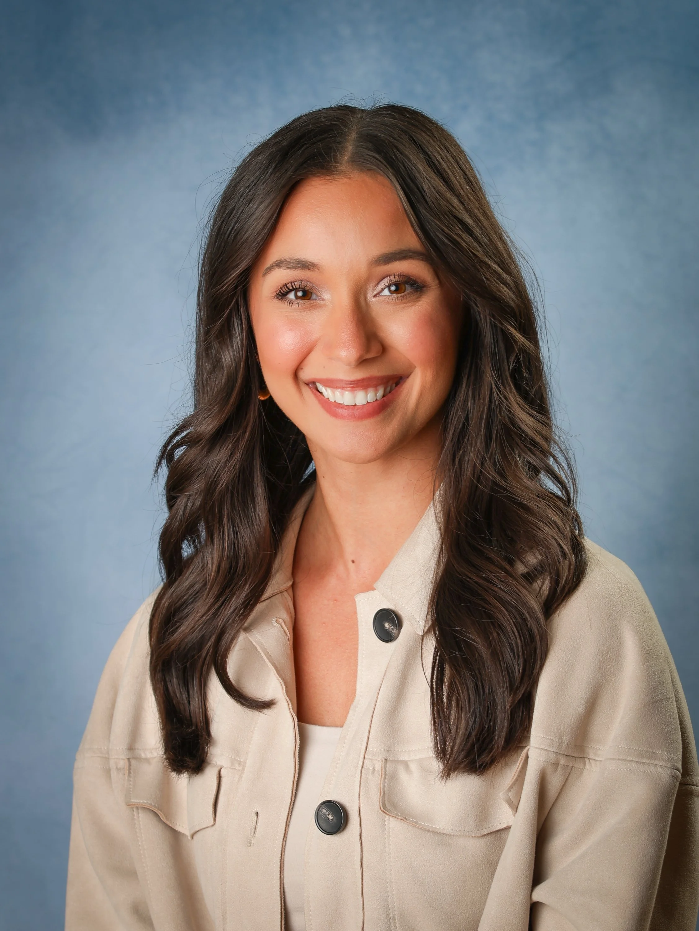 Smiling woman with long wavy brown hair, wearing a beige jacket and a white top, against a blue background.