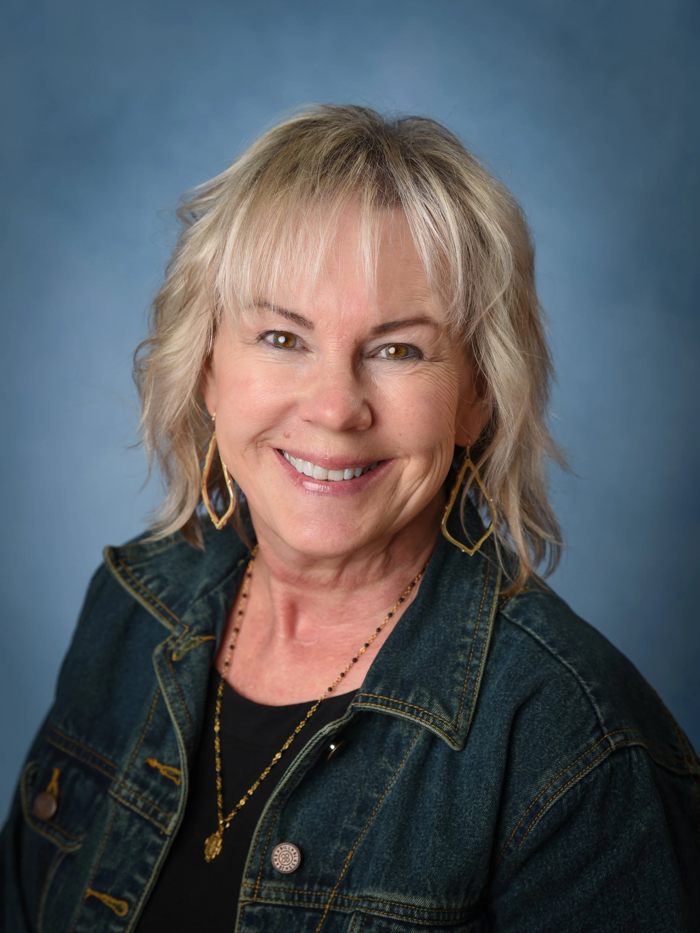 Portrait of a smiling woman with short blonde hair, wearing earrings, a necklace, and a denim jacket, against a blue background.