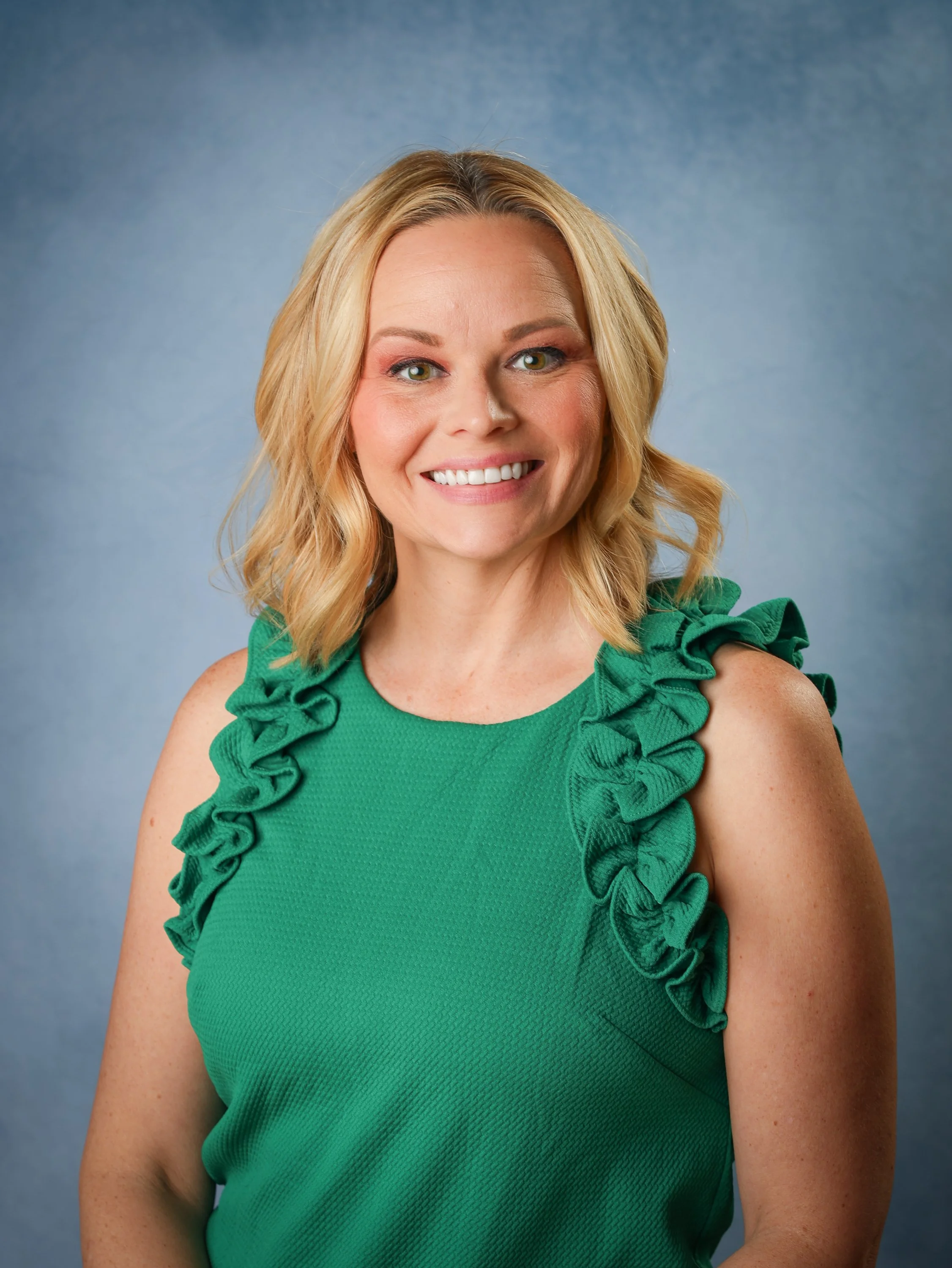 A smiling blonde woman wearing a green sleeveless top with ruffled shoulder details, standing against a blue textured background.