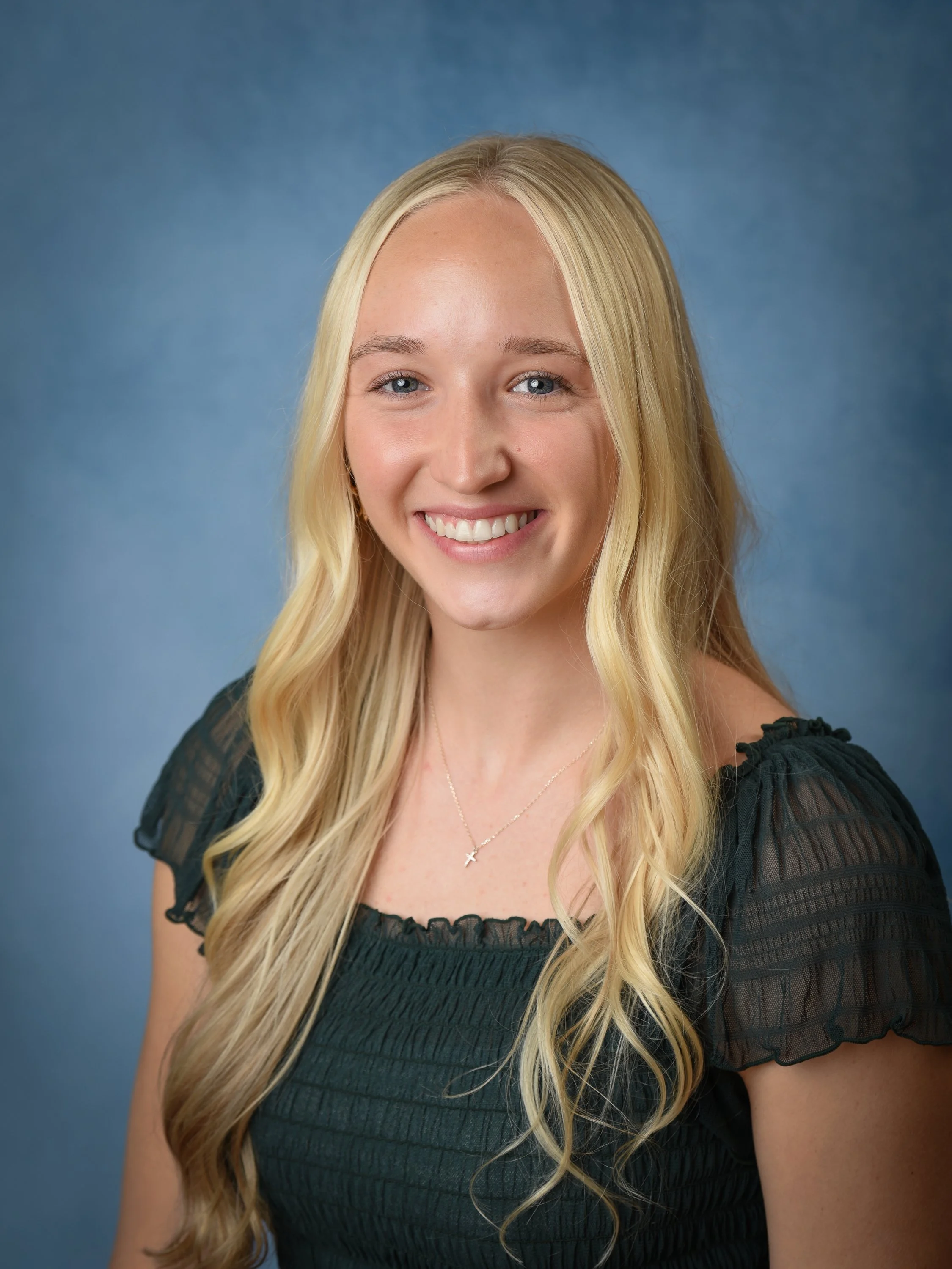 Young woman with long blonde hair, smiling, wearing a black top with ruffled sleeves and a necklace with a cross pendant, against a blue background.