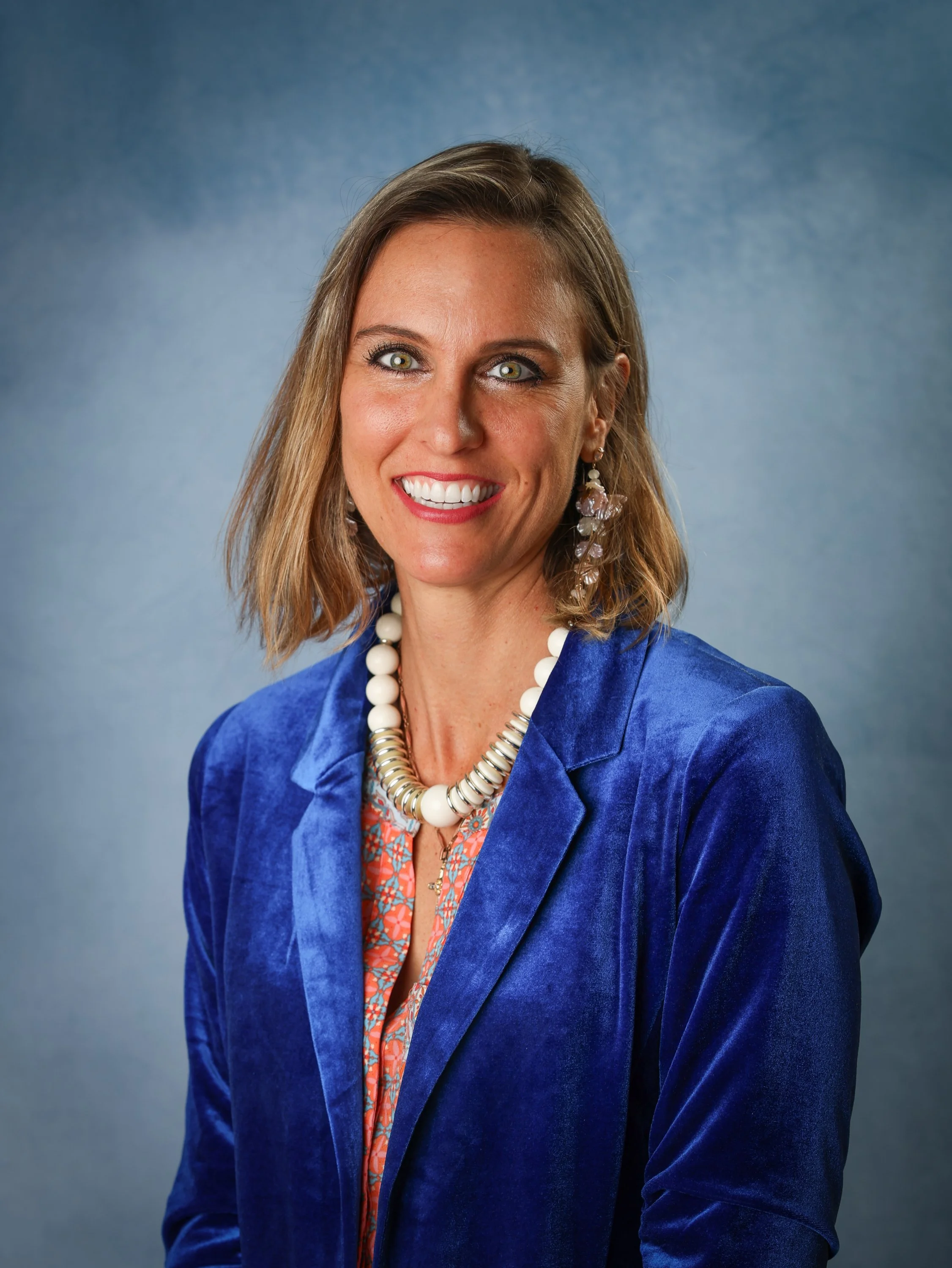 A woman smiling, wearing a blue blazer, colorful blouse, large jewelry, and earrings, against a blue background.