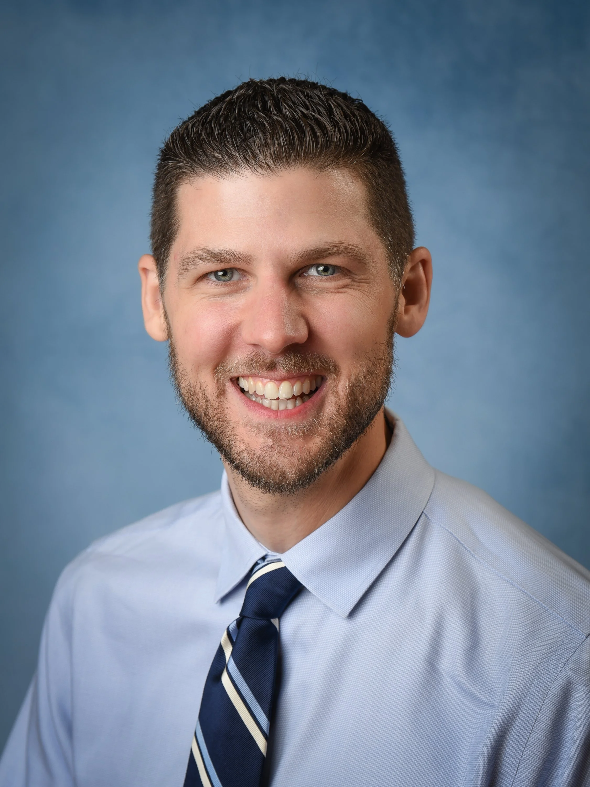 Professional headshot of a man with a beard and short brown hair, wearing a light blue shirt and a dark striped tie, smiling against a blue background.