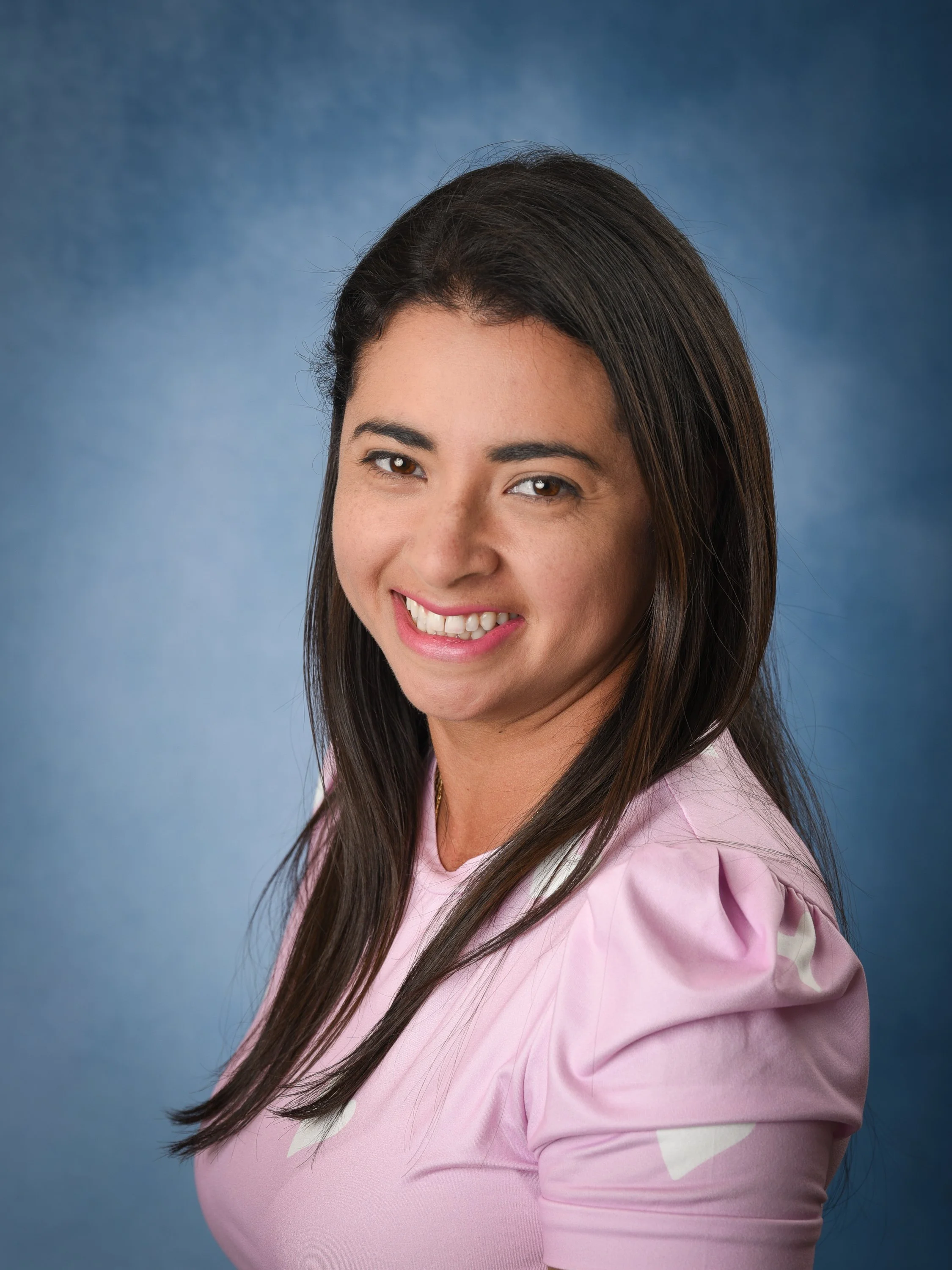 Smiling woman with shoulder-length dark brown hair and dark eyes, wearing a pink blouse against a blue background.