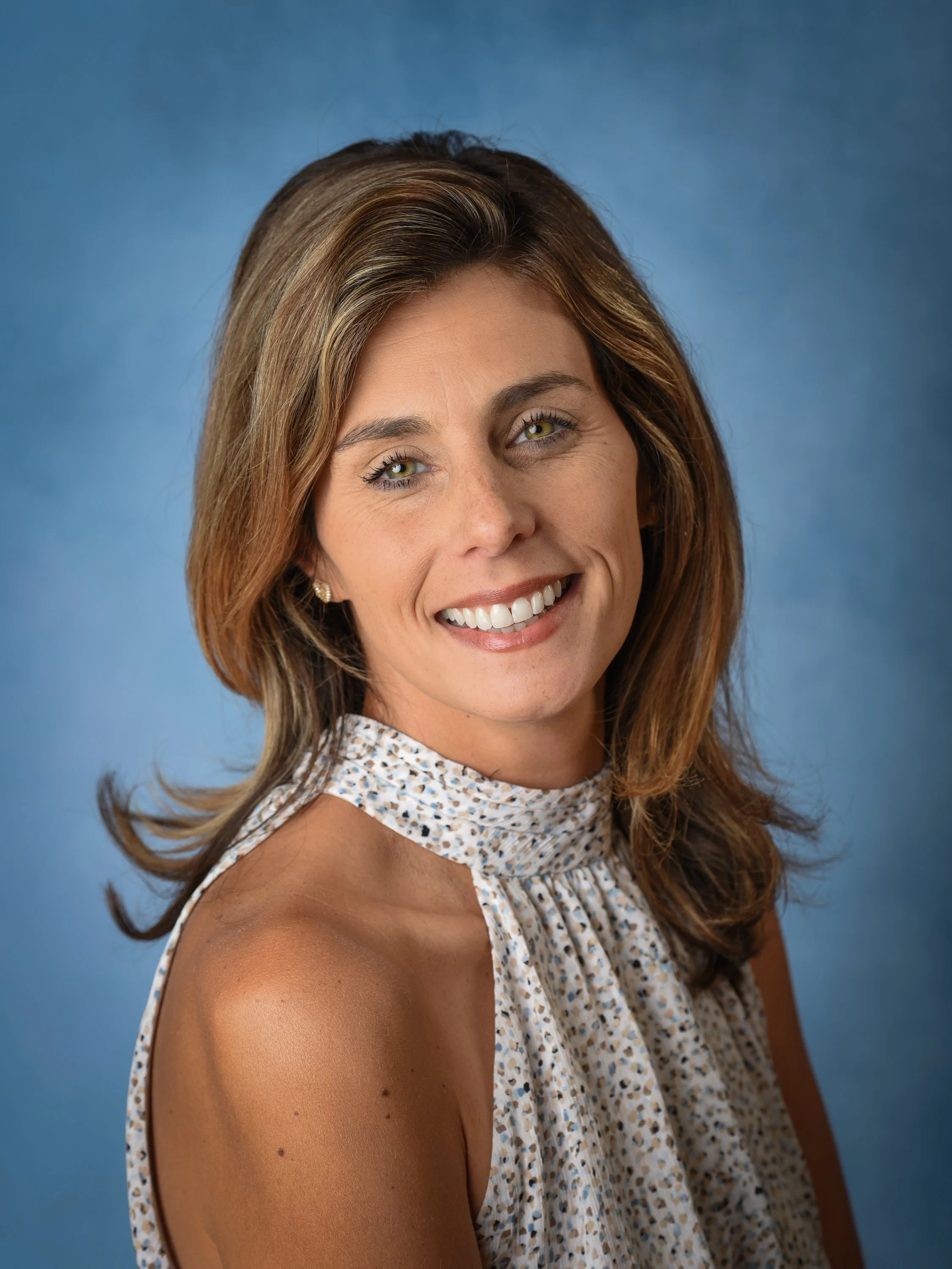 A smiling woman with shoulder-length brown hair, wearing a sleeveless patterned blouse, posing against a blue background.