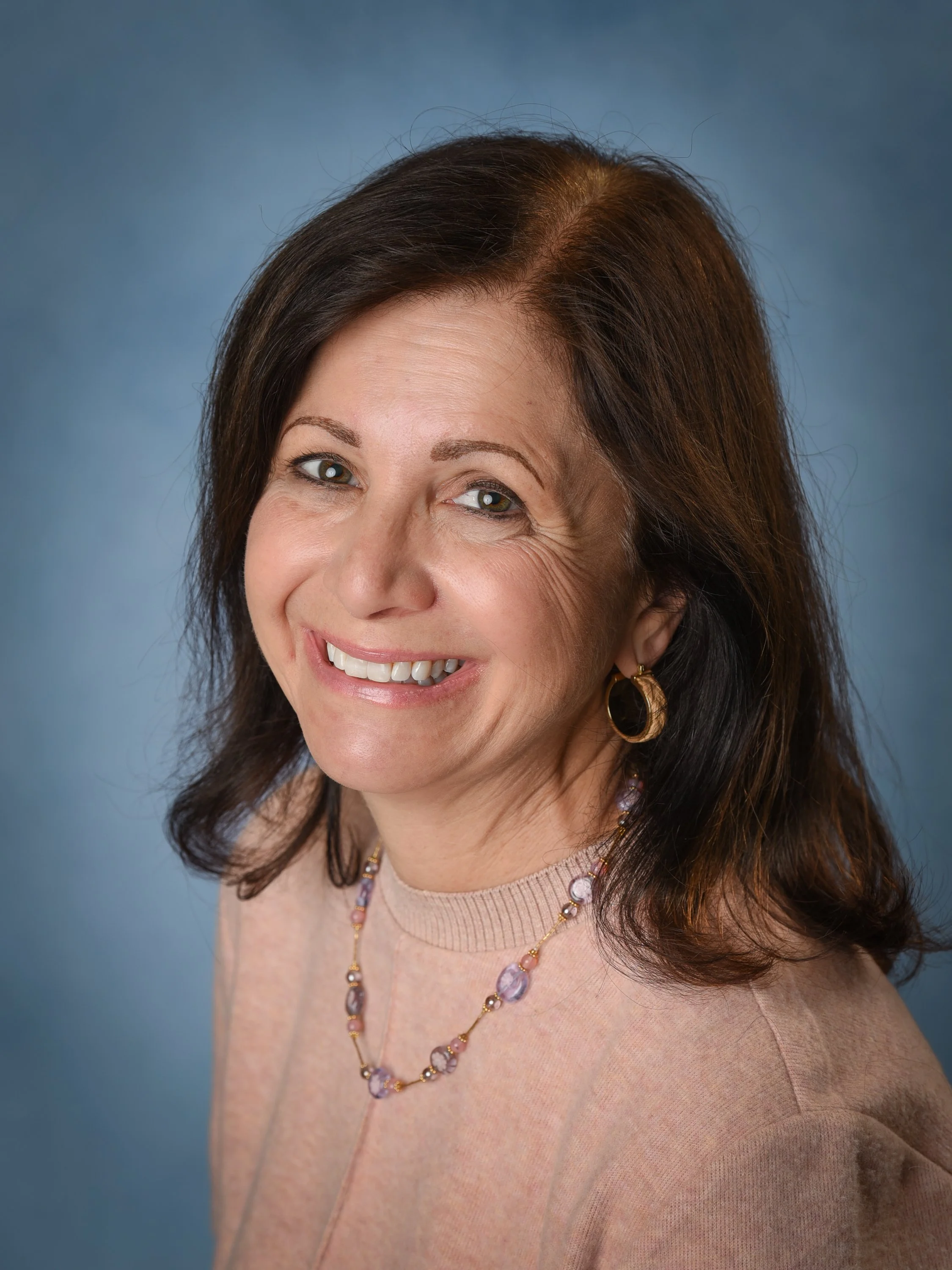 Portrait photo of a smiling woman with shoulder-length brown hair, wearing a beaded necklace and gold hoop earrings against a blue background