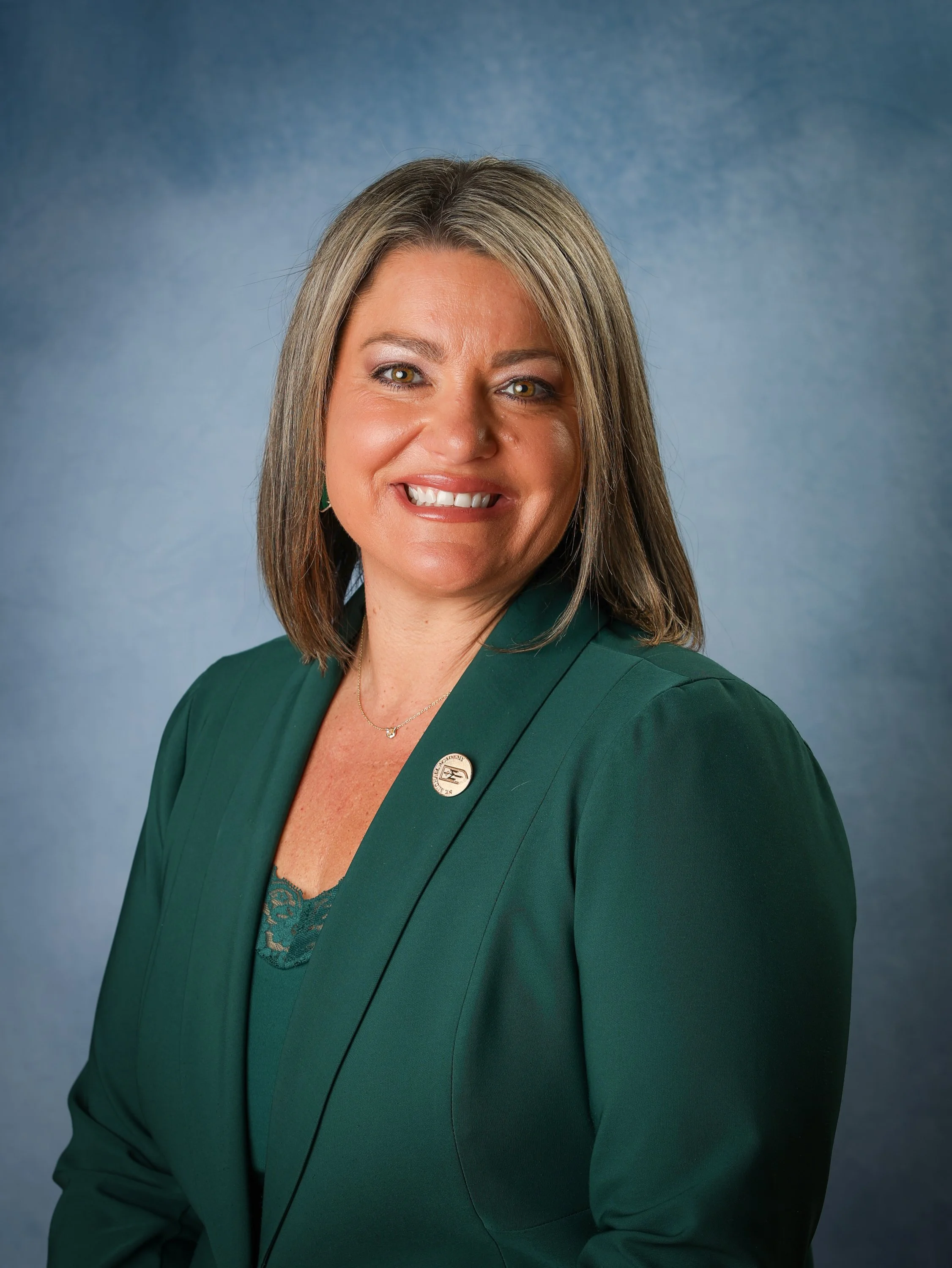 Headshot of a woman with shoulder-length brown hair, smiling, wearing a green blazer with a pin and a necklace, against a blue background.