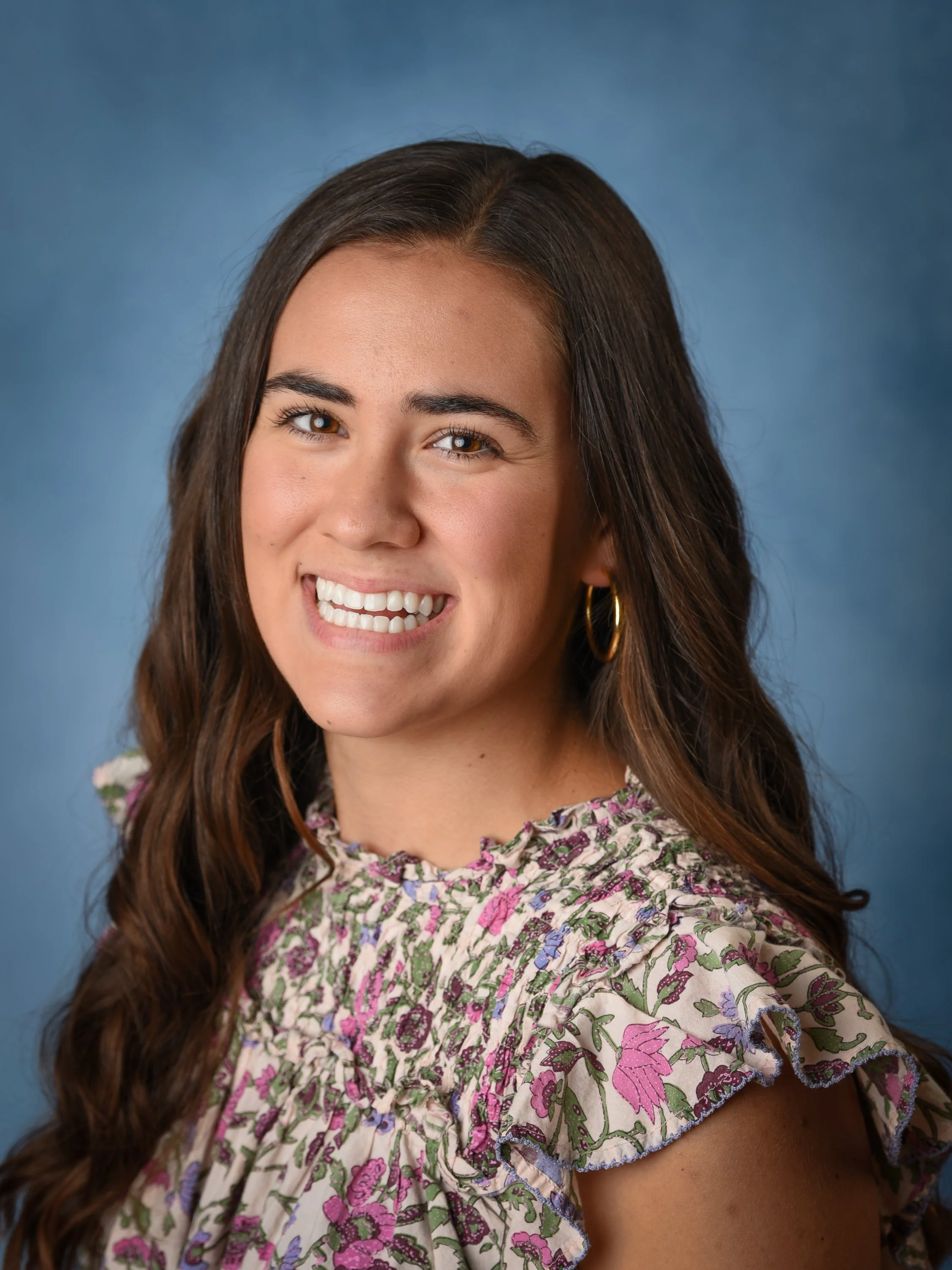 Close-up portrait of a young woman with long wavy brown hair, wearing a floral ruffled blouse and gold hoop earrings, smiling against a blue background.