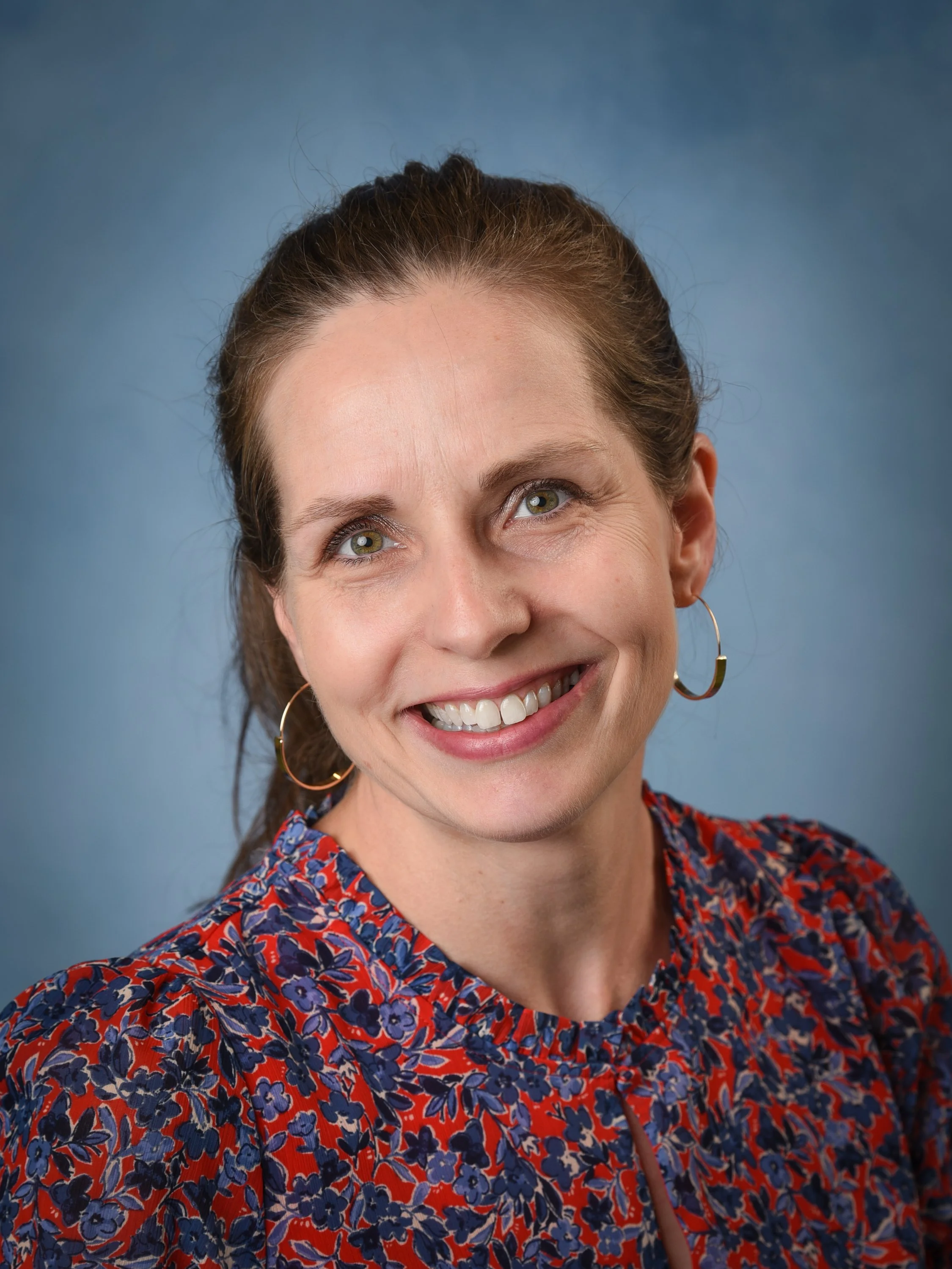 Portrait of a smiling woman with shoulder-length brown hair, green eyes, large hoop earrings, and a floral-patterned top against a blue background.