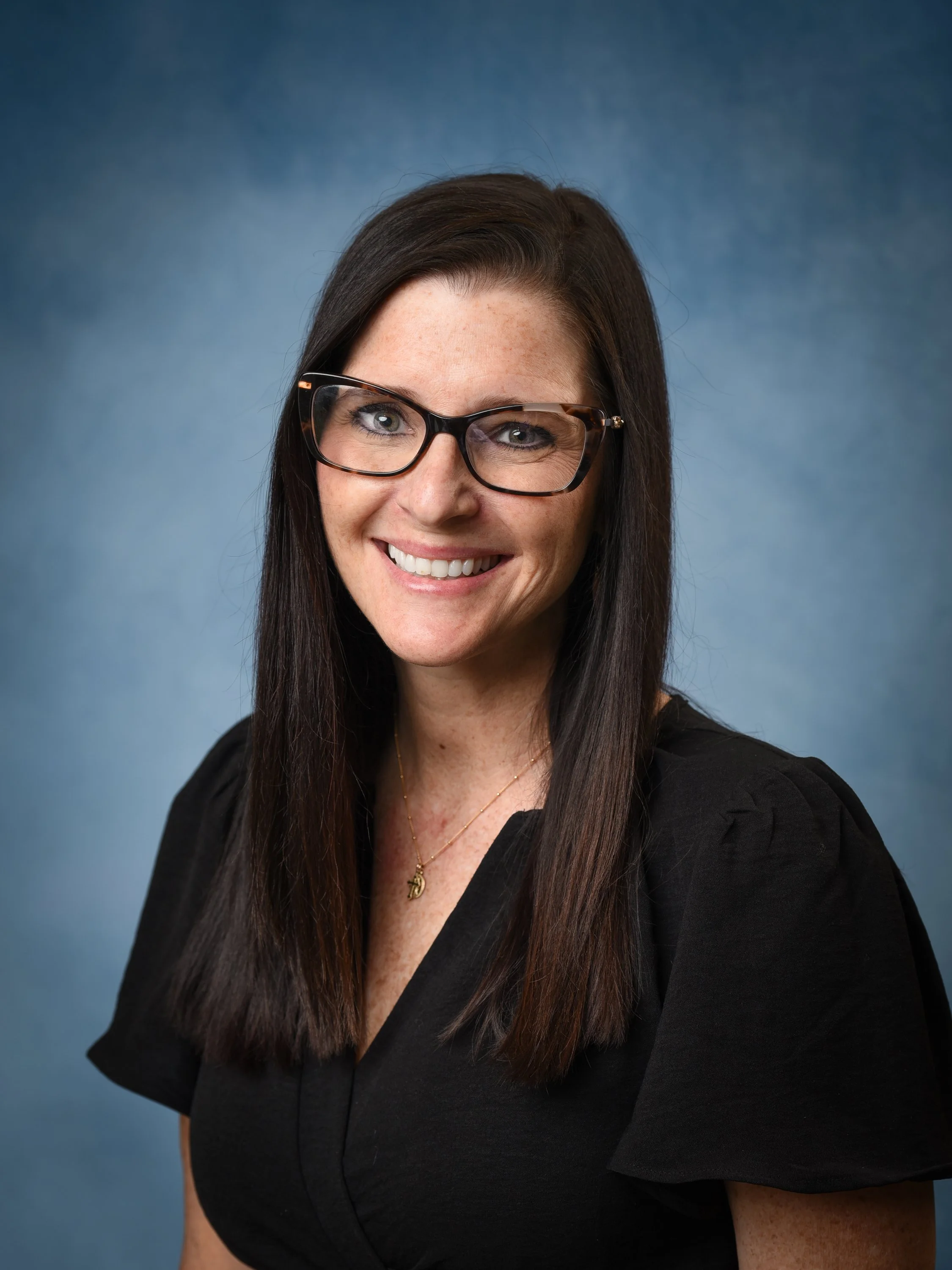 Smiling woman with long dark brown hair and dark-framed eyeglasses, wearing a black top with a necklace against a blue background.
