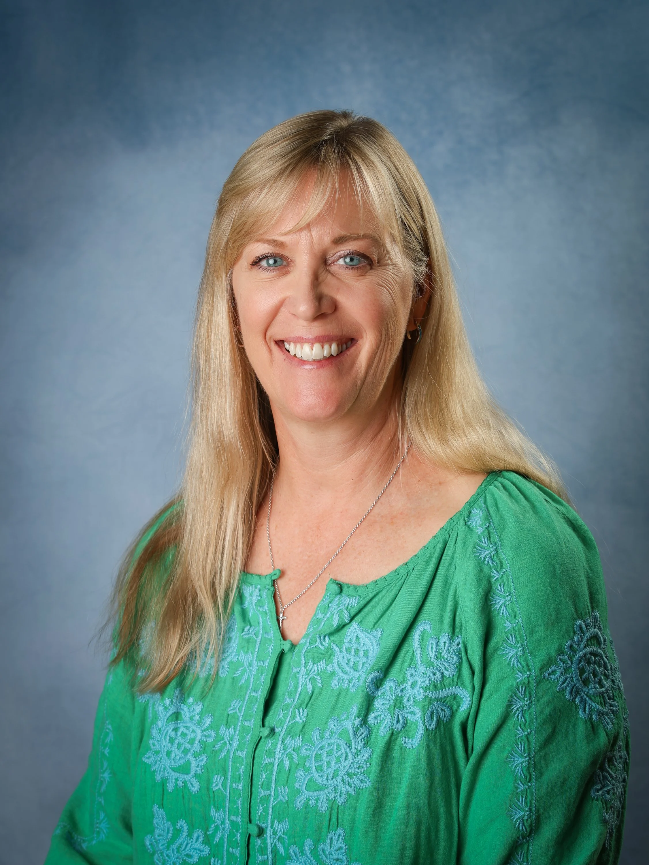 Portrait of a smiling woman with blonde hair, wearing a green embroidered blouse, silver necklace, and earrings, against a blue gradient background.