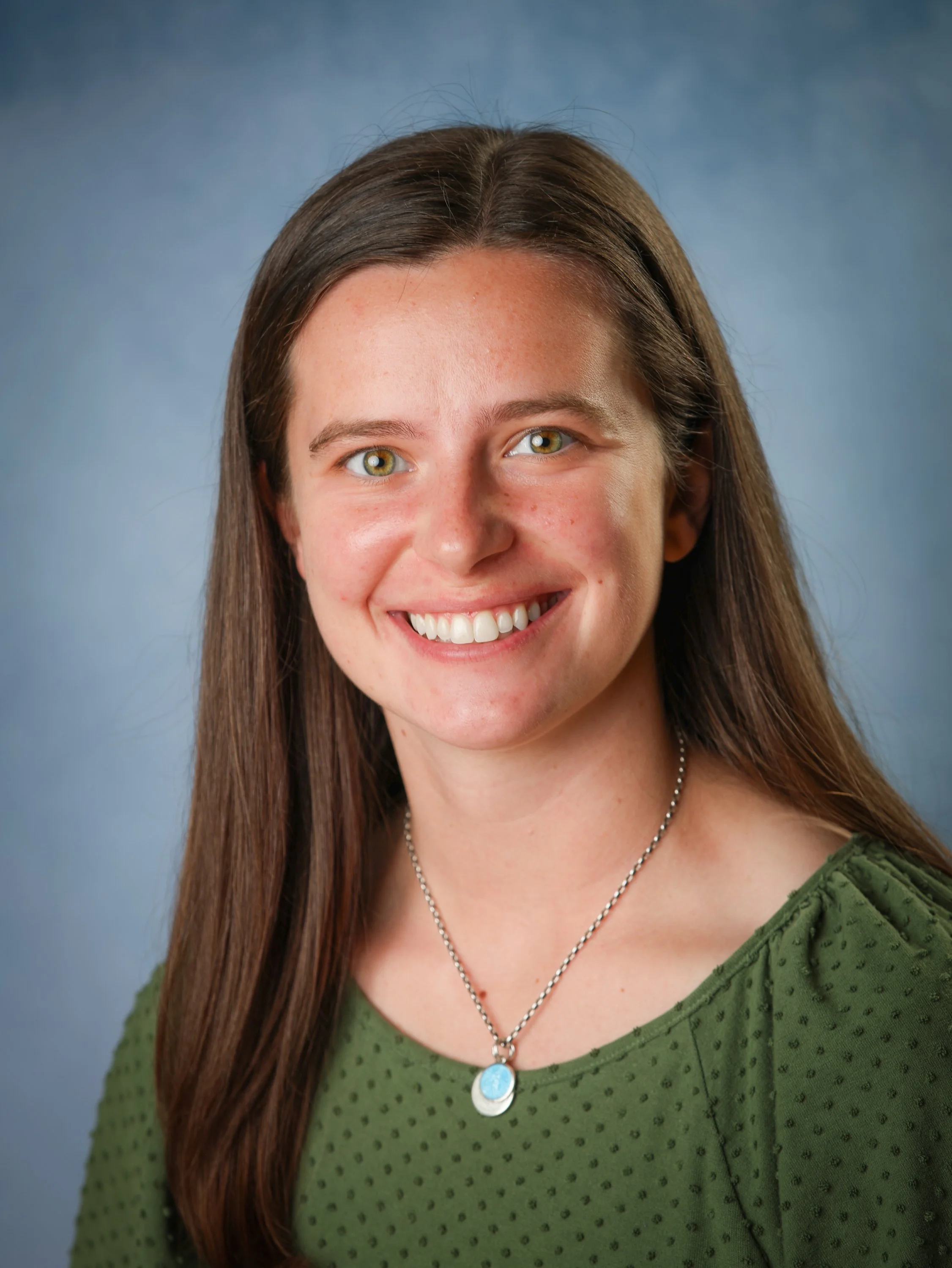 A young woman with long brown hair, green eyes, and a bright smile, wearing a green dotted top and a necklace with a turquoise pendant, against a blue-gray background.