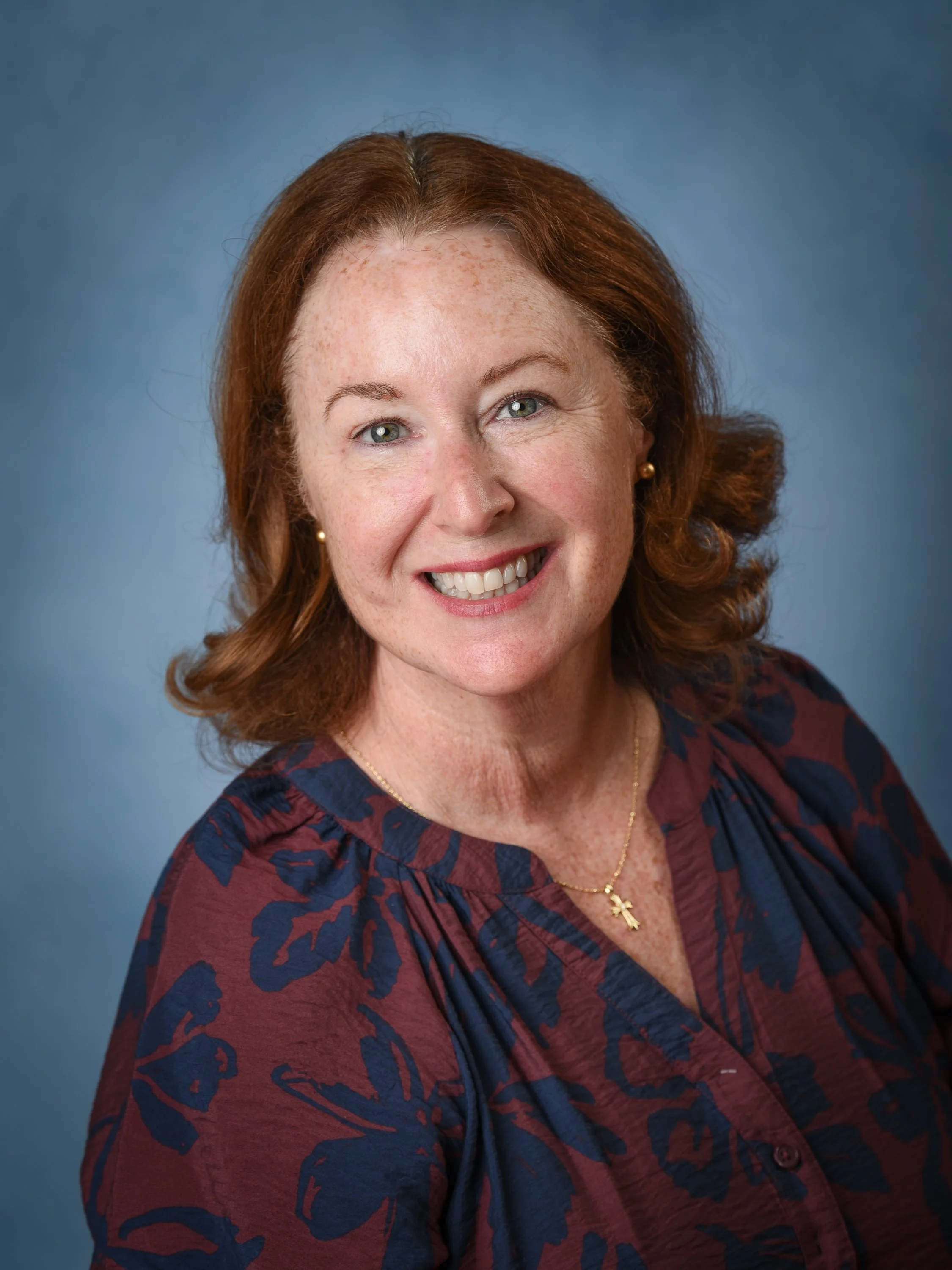 Portrait of a smiling woman with red hair, fair skin, wearing a dark floral blouse, a gold cross necklace, and pearl earrings, against a solid blue background.