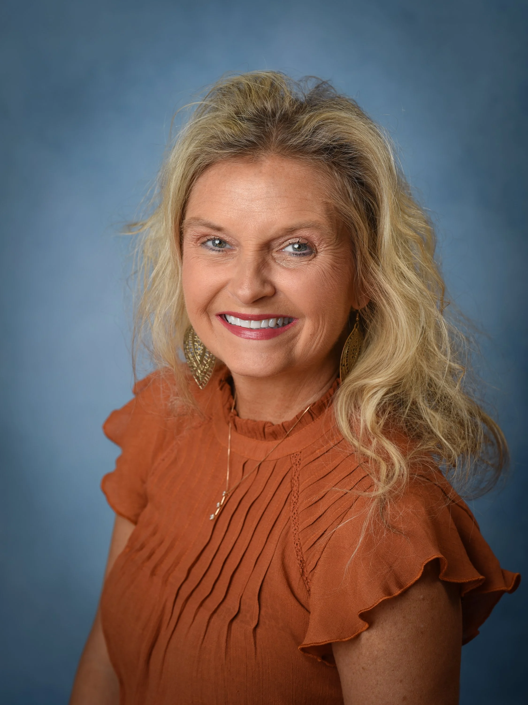 A woman with blonde hair and blue eyes, smiling, dressed in an orange top with ruffled sleeves, wearing large earrings and a necklace, against a blue background.