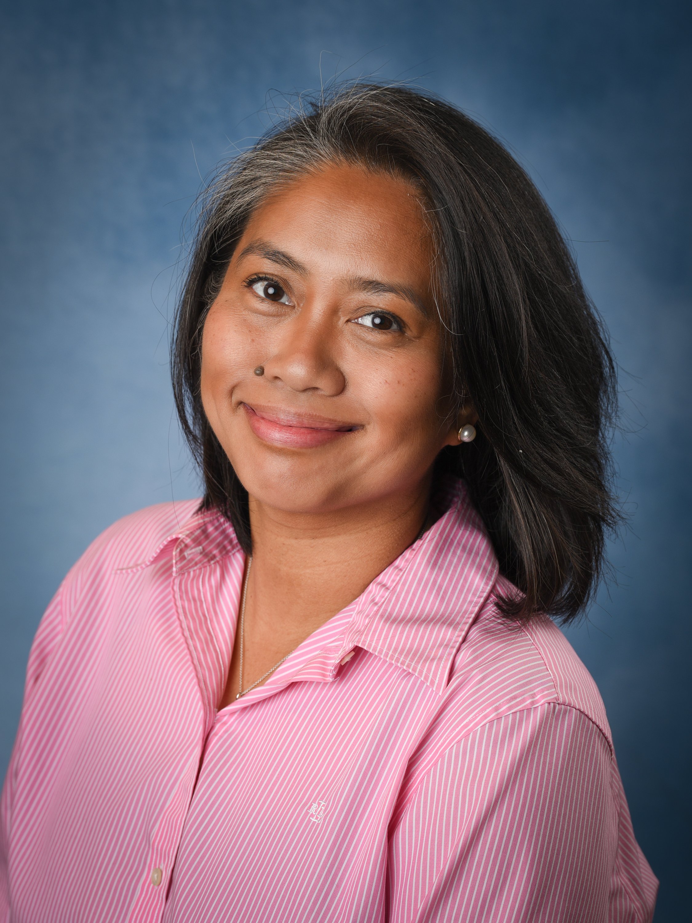 smiling woman with shoulder-length graying brown hair and pearl earrings, wearing a pink striped shirt against a blue background.