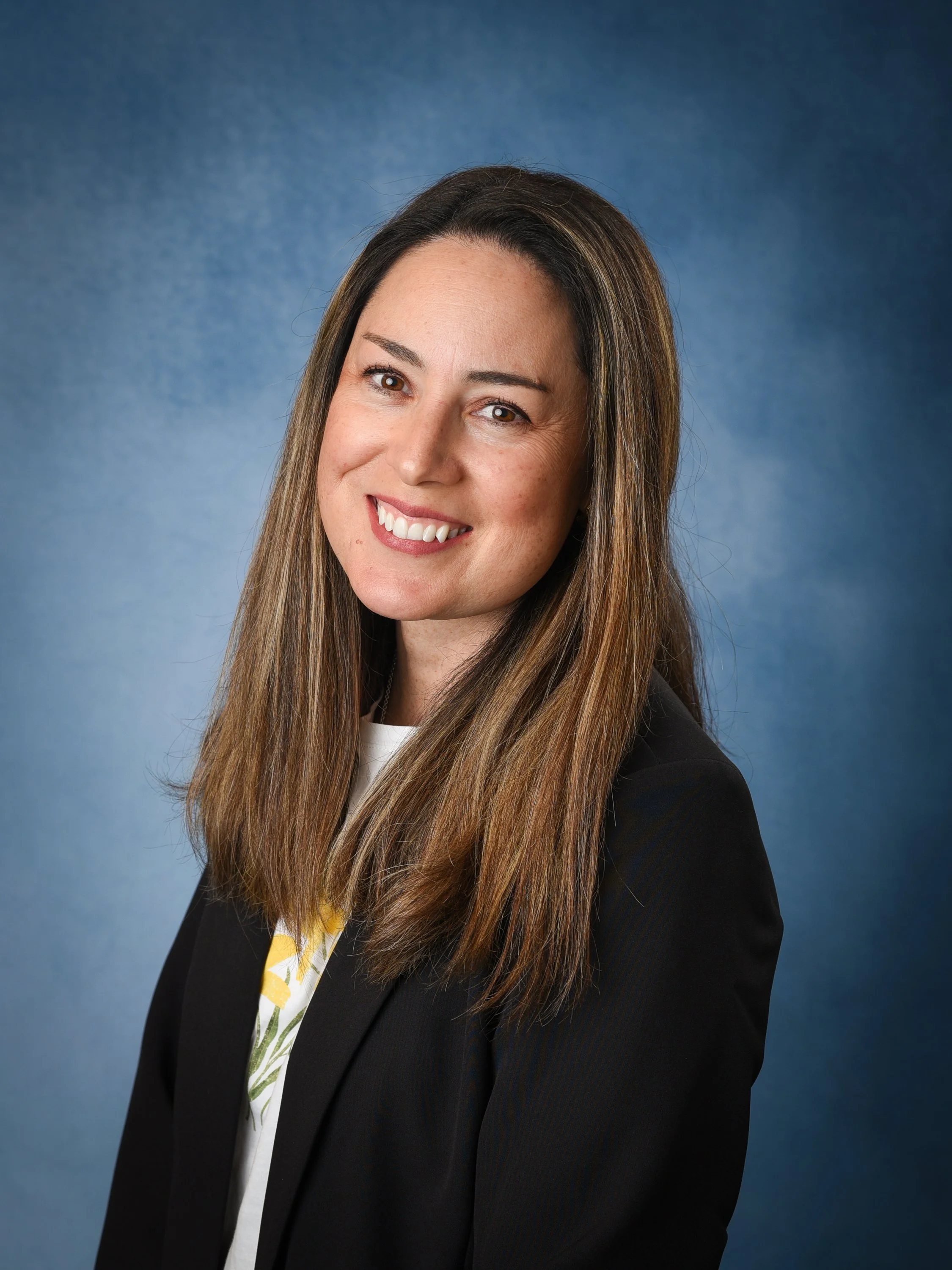 Professional headshot of a smiling woman with long brown hair, wearing a blazer and white top against a blue background.