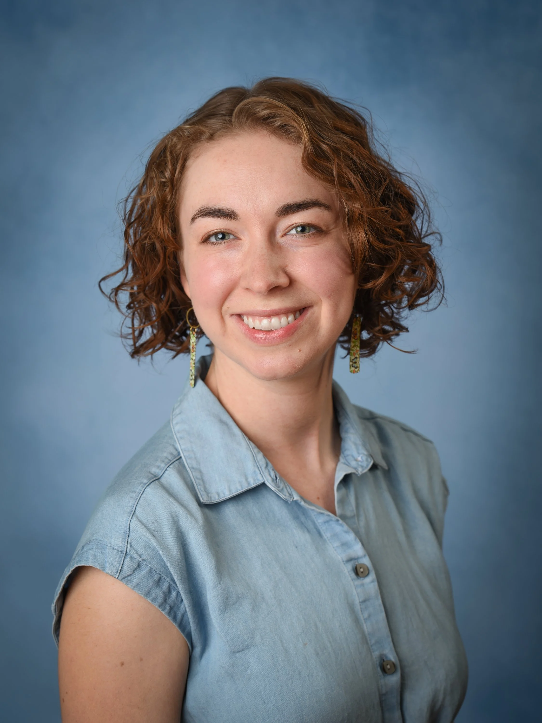 Close-up portrait of a smiling woman with curly red hair wearing a light blue denim shirt and dangling earrings against a blue background.