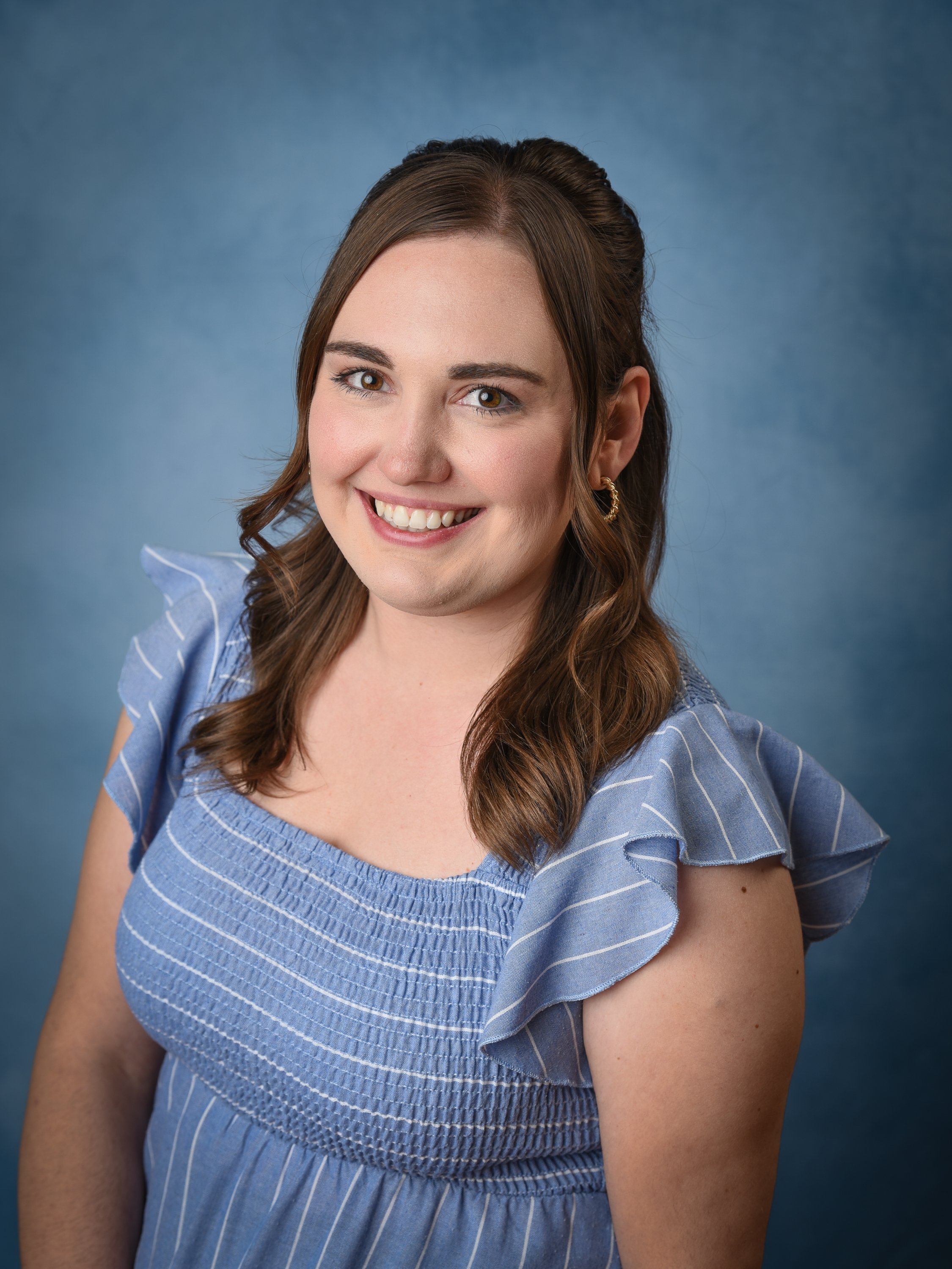 A woman with brown hair smiling, wearing a blue dress with ruffled sleeves, posed against a blue gradient background.