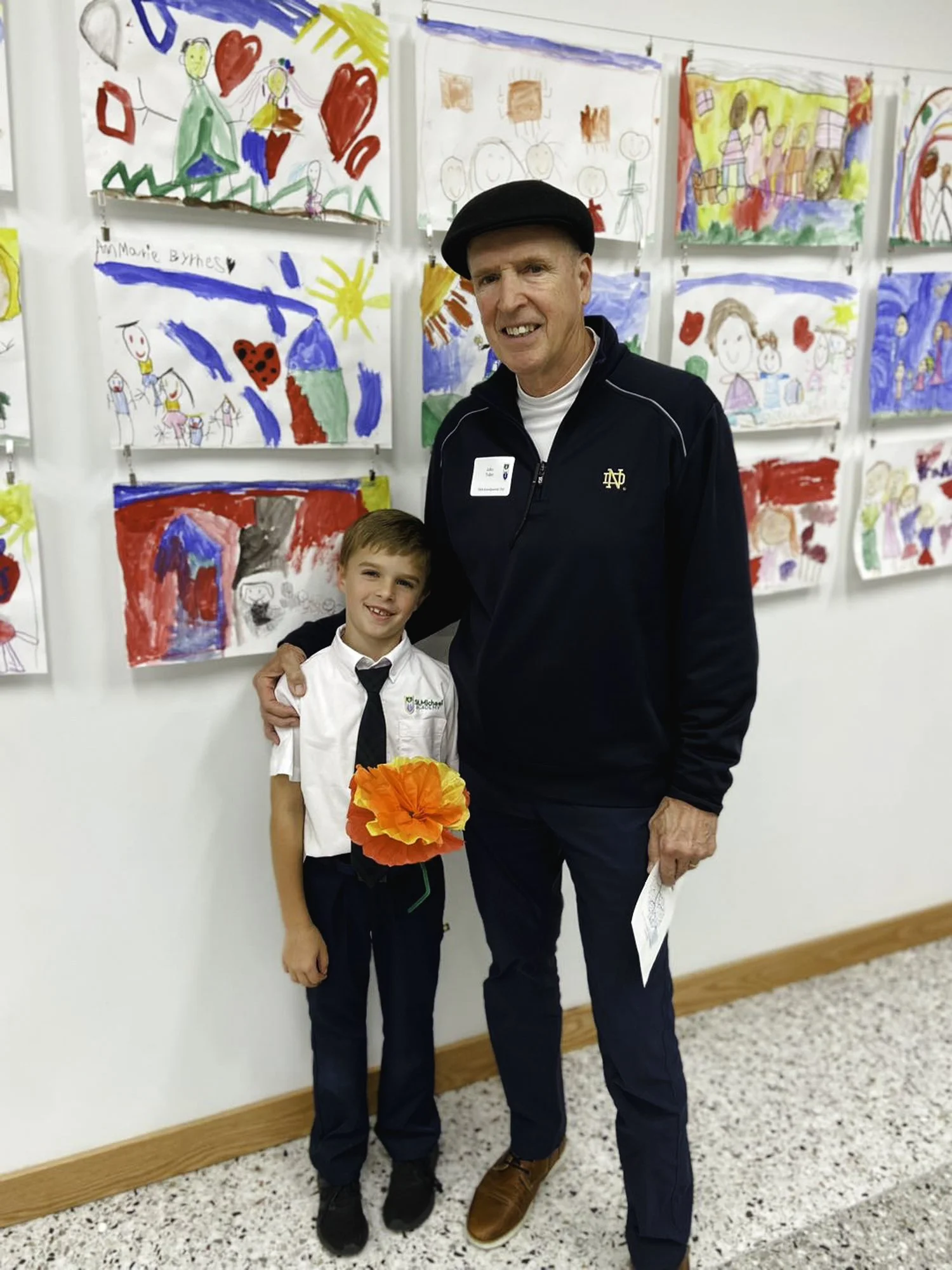 A young boy and an adult man smile and pose together in front of a wall covered with children's colorful drawings. The boy is holding a large, bright orange paper flower.