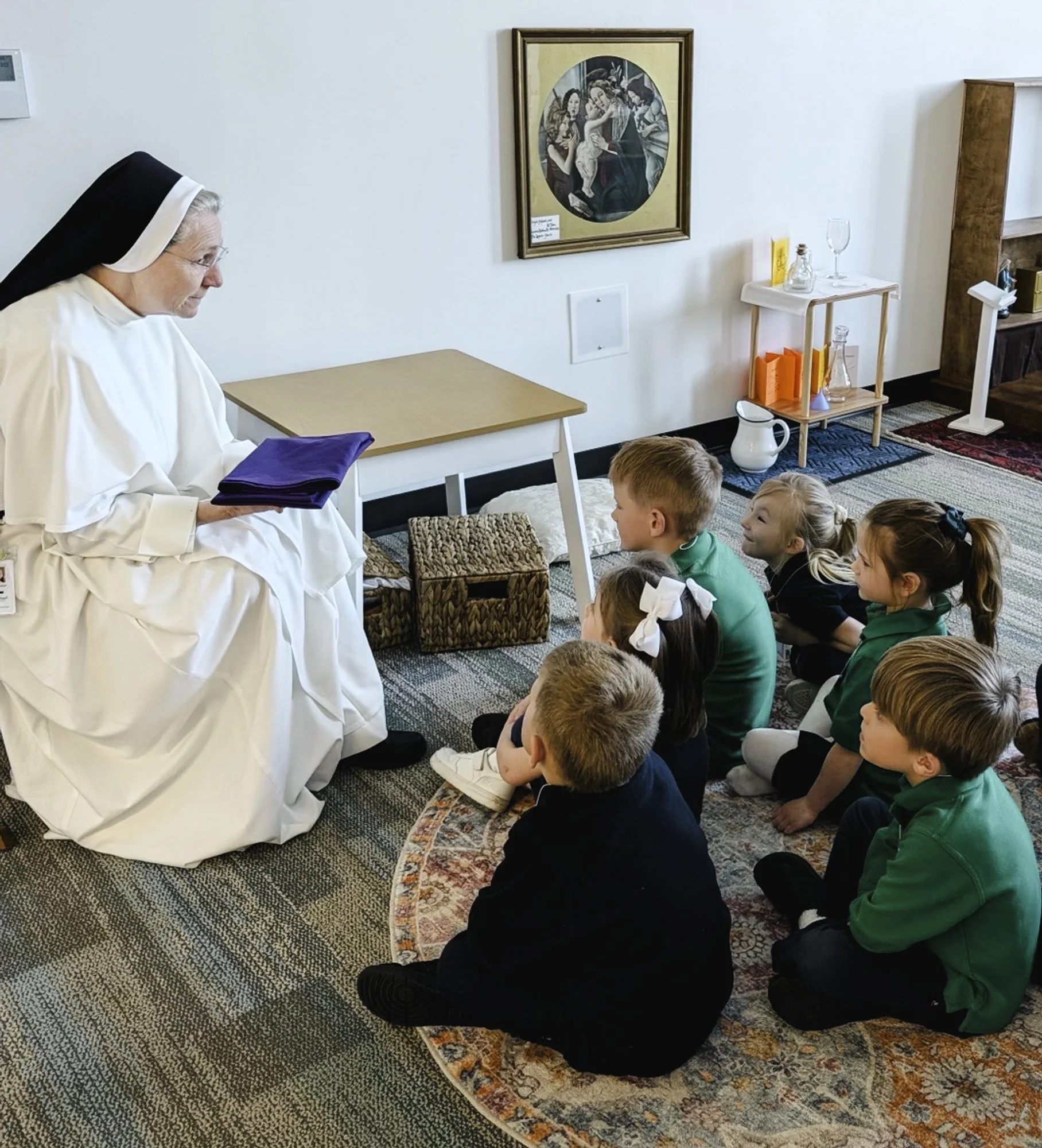 A nun in white habit sitting in front of a group of children sitting on a patterned rug, listening to her in a room with religious artwork and books on shelves.
