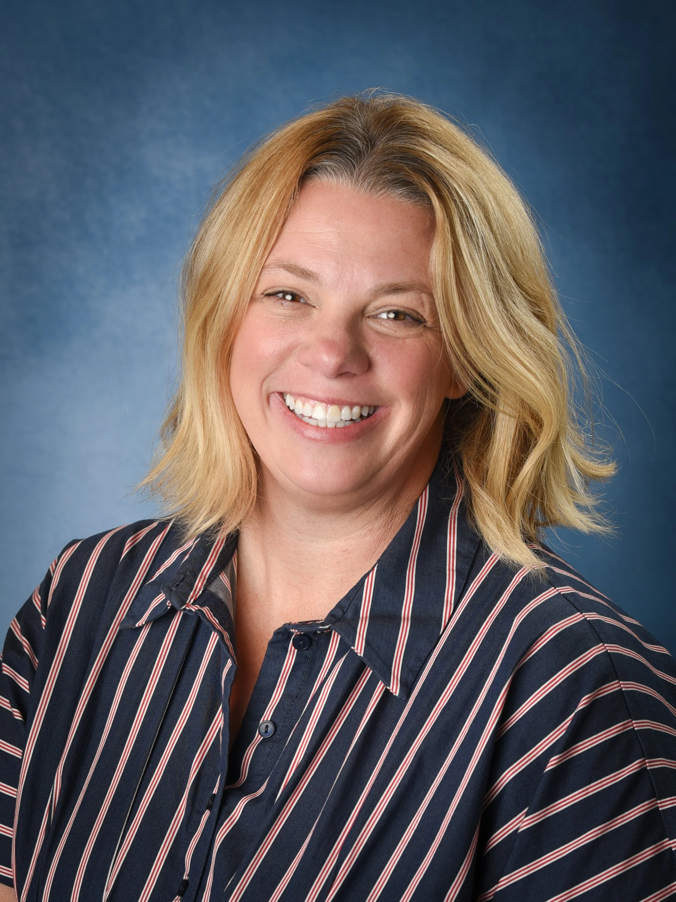 Smiling woman with wavy blonde hair, wearing a striped shirt collar against a blue background.