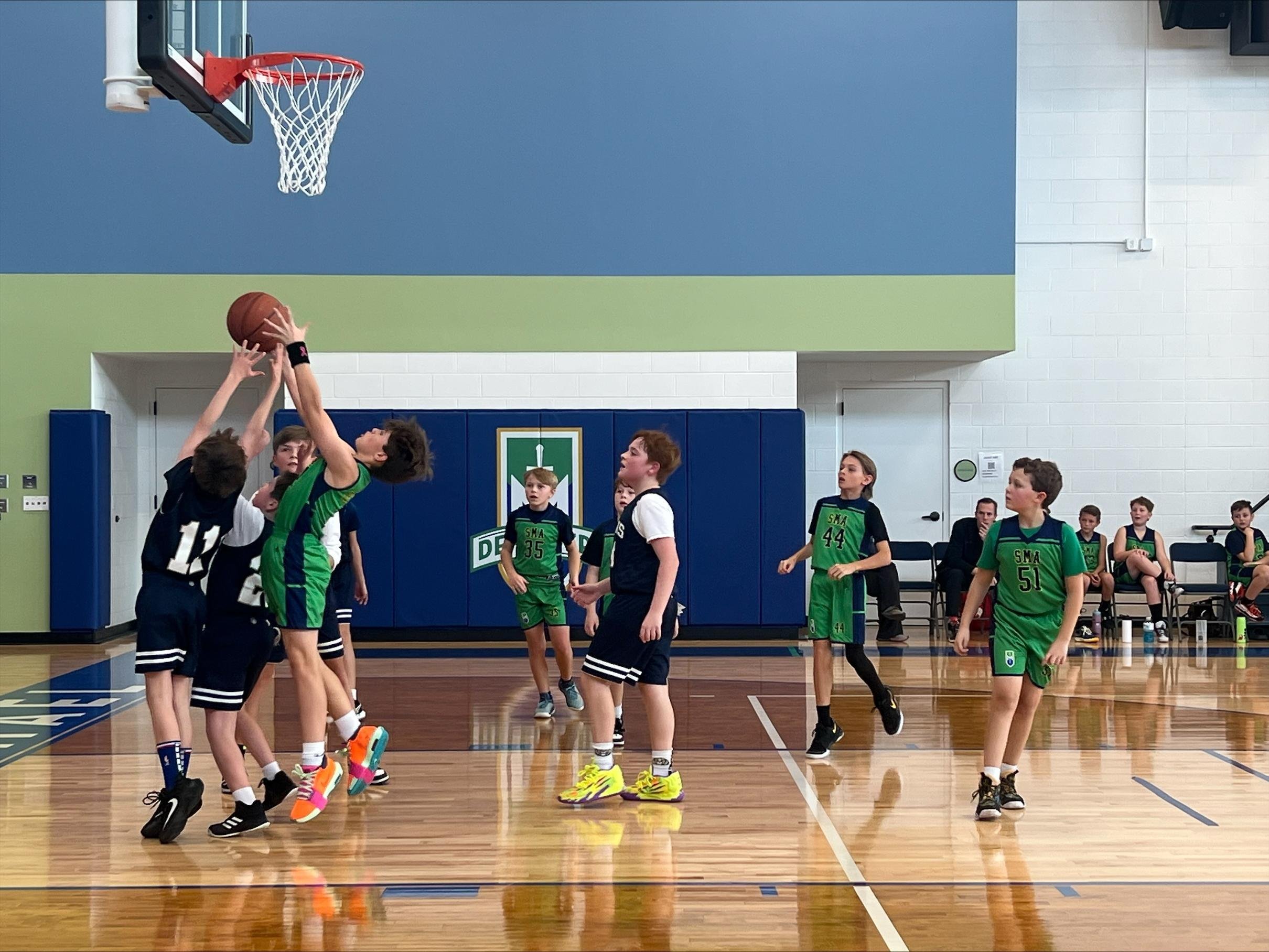Group of children playing basketball in an indoor gym, with some children attempting a shot and others watching.