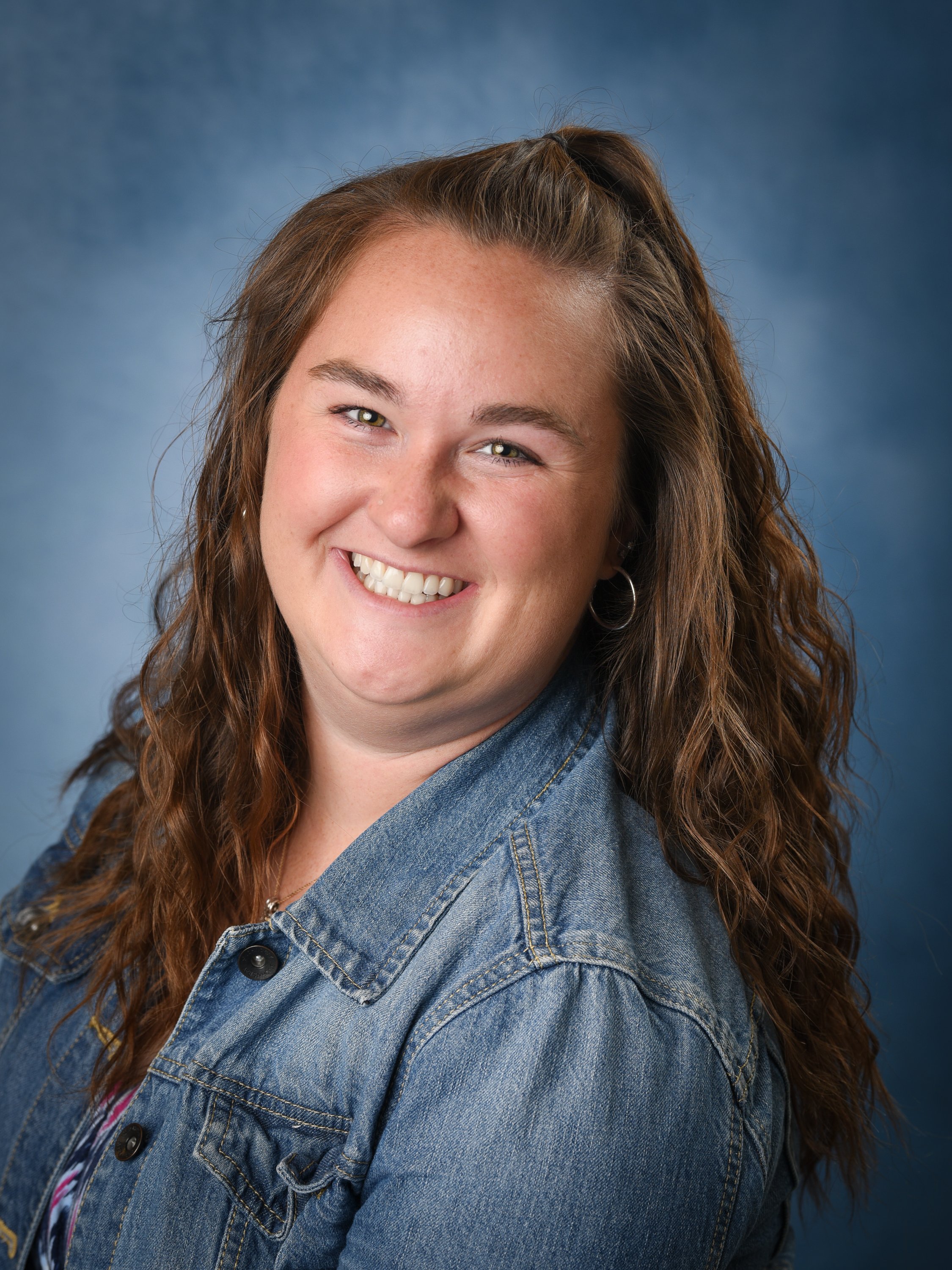 A smiling woman with long curly red hair, wearing a denim jacket, against a blue background.