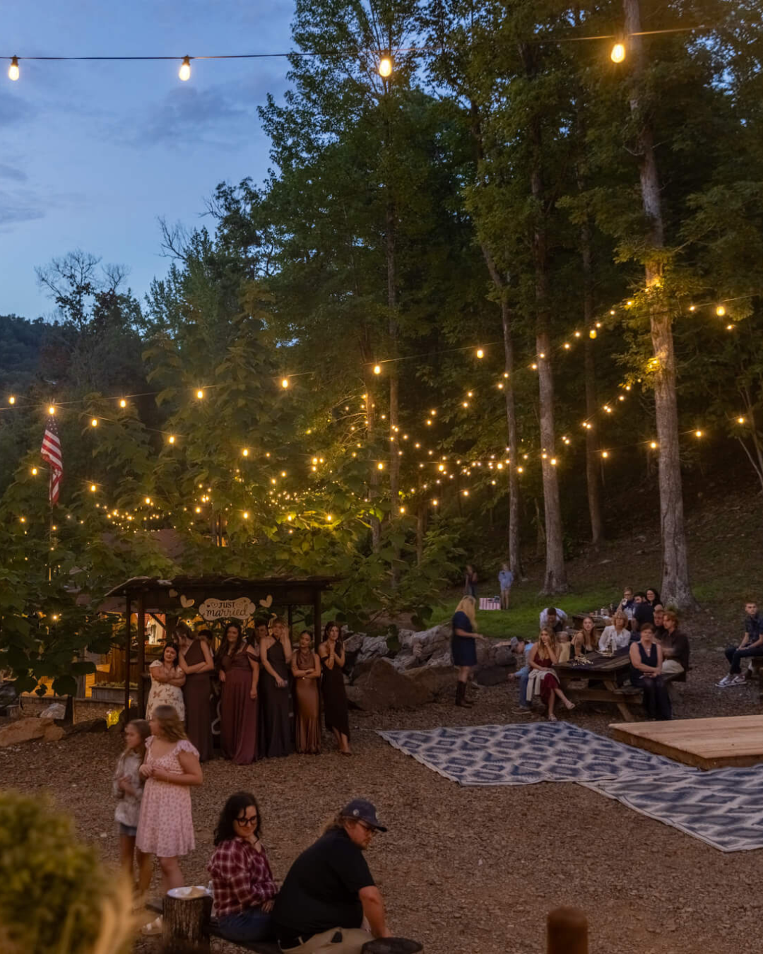 An outdoor wedding reception at dusk with string lights overhead, a wooded background, and a group of people gathered, some sitting at picnic tables, others standing or walking, with a