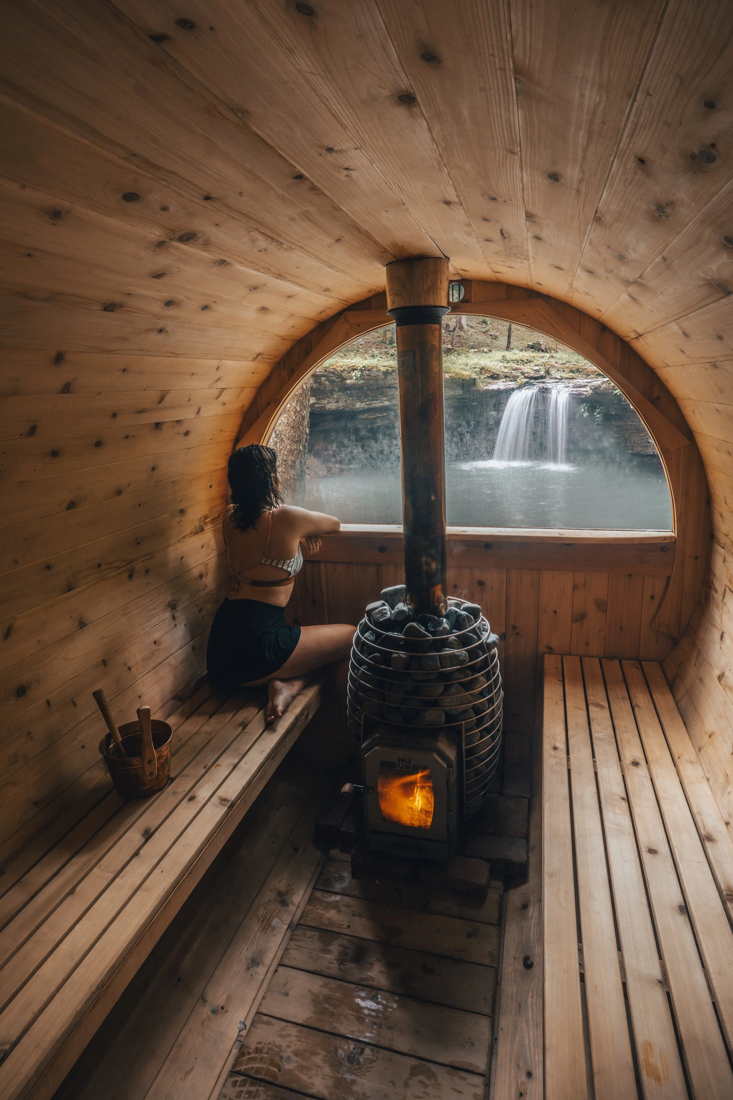 A person sitting inside a wooden sauna, looking out the window at a waterfall and natural landscape, with a stove and rocks in the center, and a bucket and ladle nearby.