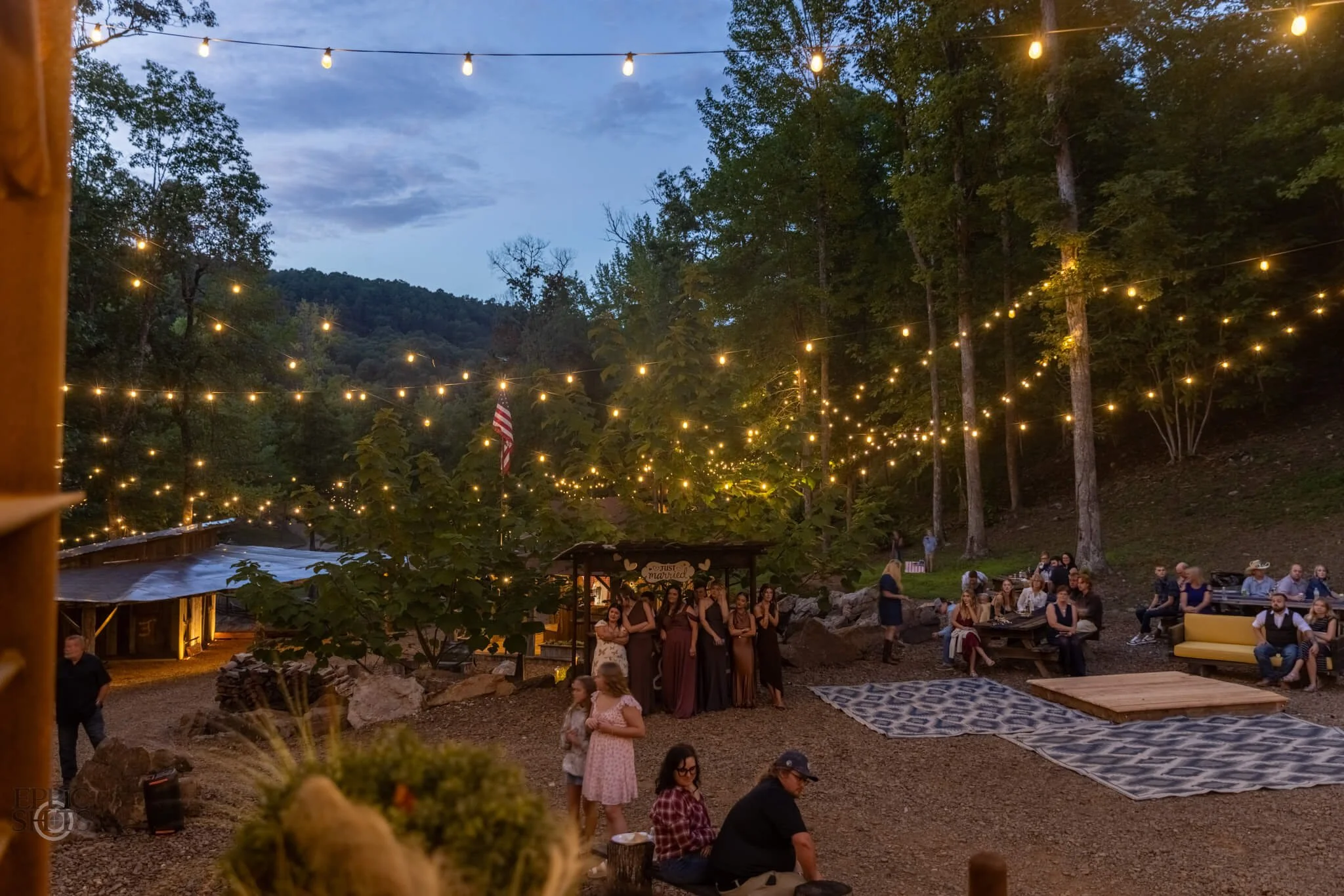 Outdoor gathering with string lights illuminating a wooded area during dusk, with people standing and sitting around tables, some holding drinks, and an American flag in the background.