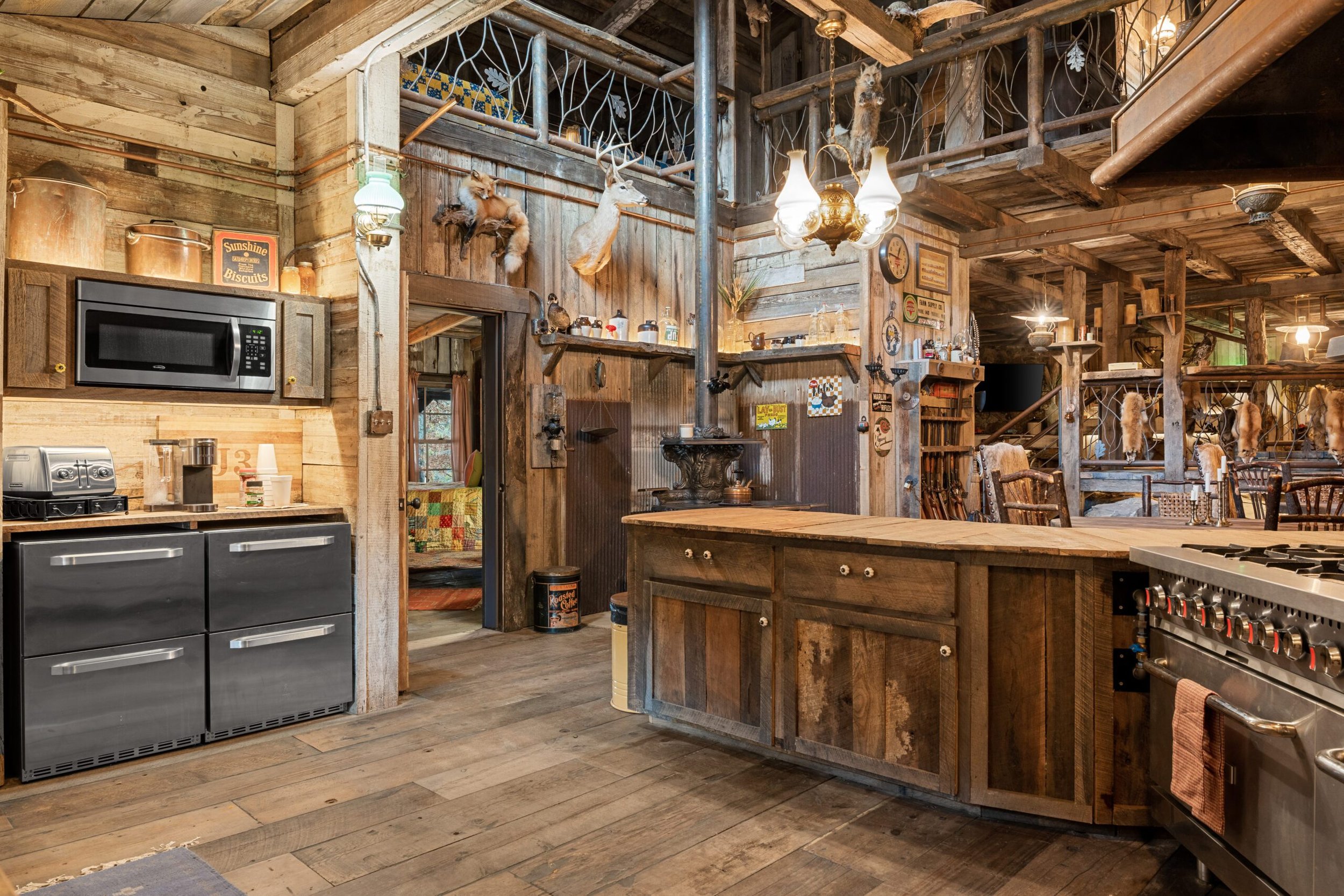 Rustic kitchen with wooden cabinetry, a microwave, and a stovetop. Decor includes animal mounts, vintage signs, and antique kitchenware. Wooden floors and exposed beams enhance the rustic ambiance.
