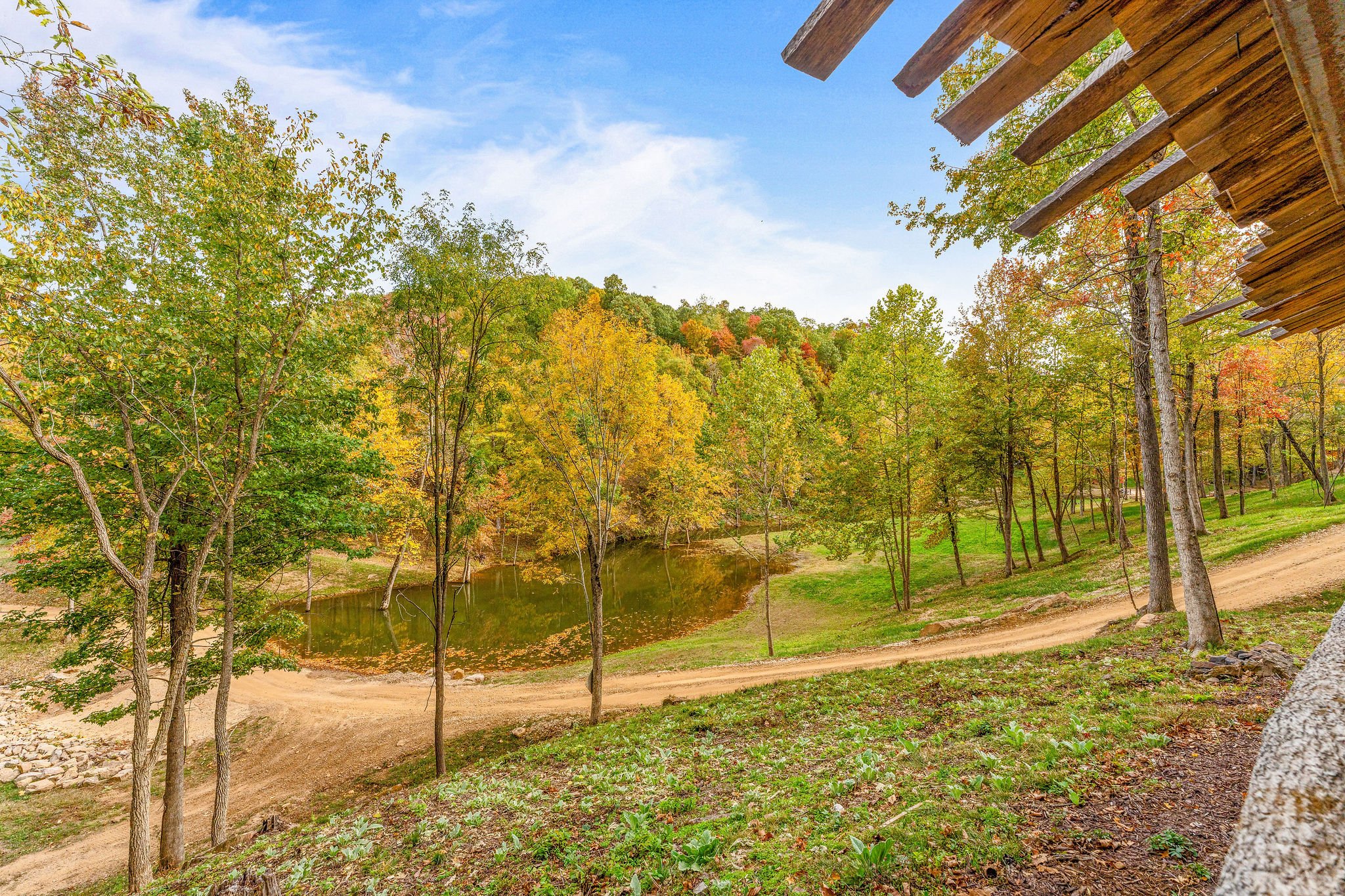 Scenic view of a forested landscape with autumn foliage, a dirt path, a small pond, and a wooden structure in the foreground.